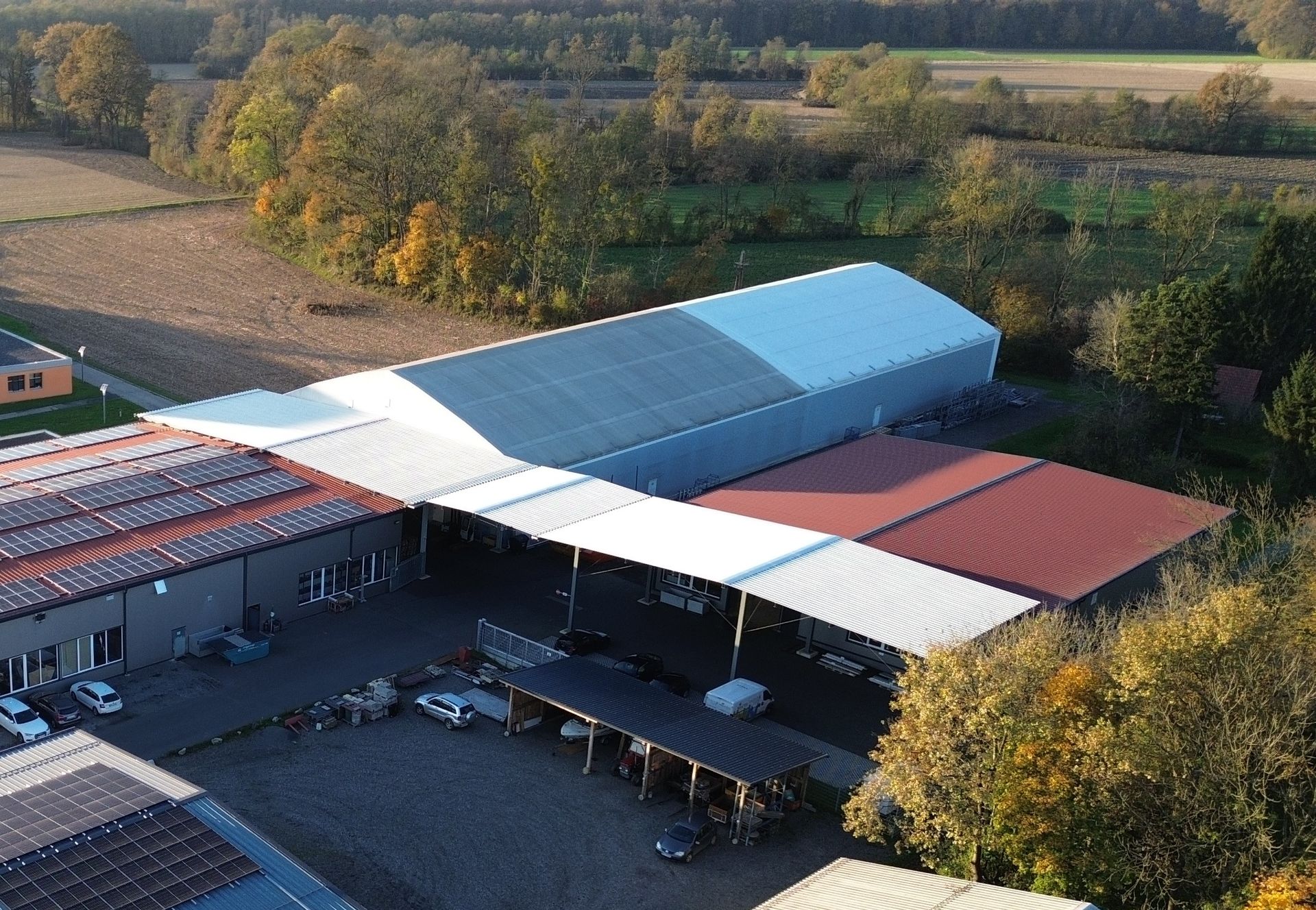 Luftaufnahme von Industriegebäuden mit silbernen und roten Dächern, umgeben von Bäumen und Feldern.