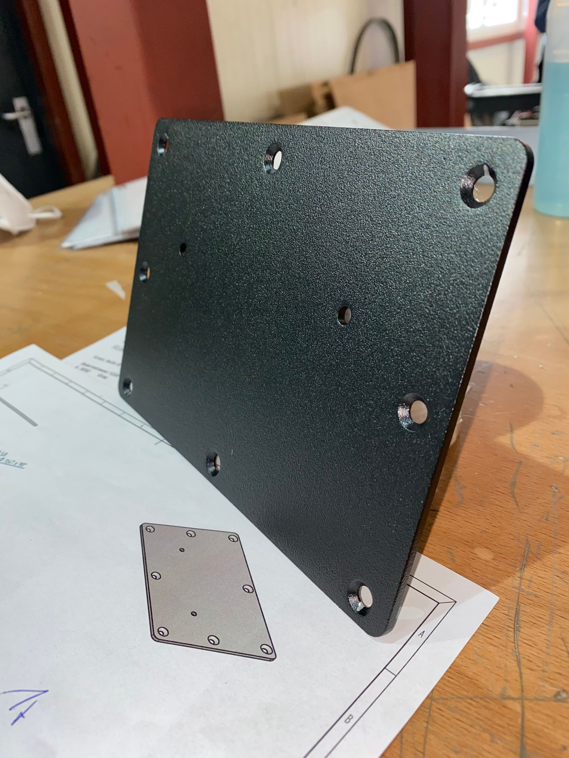 Schwarze Metall-Montageplatte mit mehreren Löchern und Nieten, auf einem Schreibtisch mit einer Bauzeichnung.