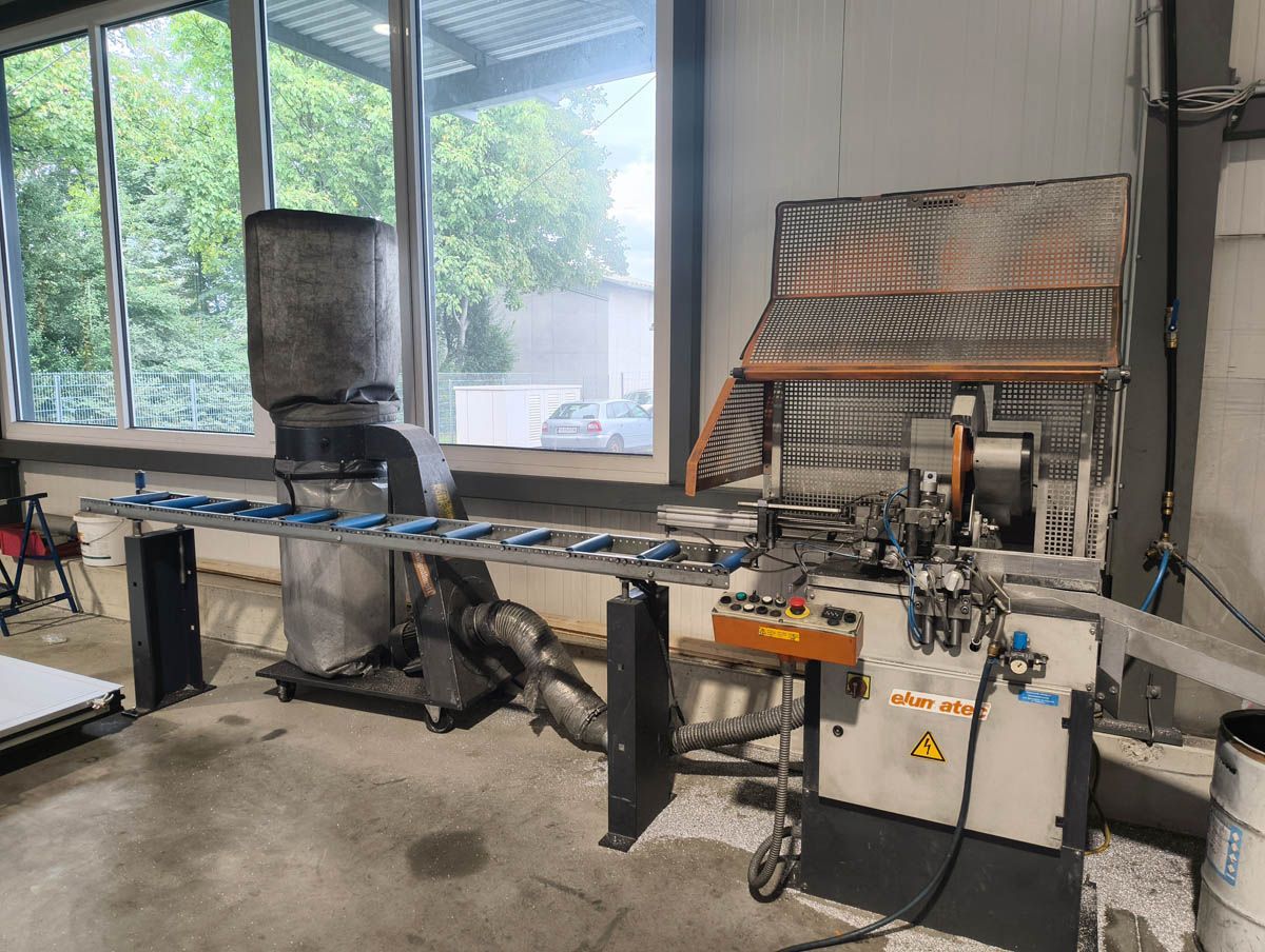Industriemaschine in einer Werkstatt, mit einem langen Förderband und einer Metallverkleidung, in der Nähe eines Fensters.