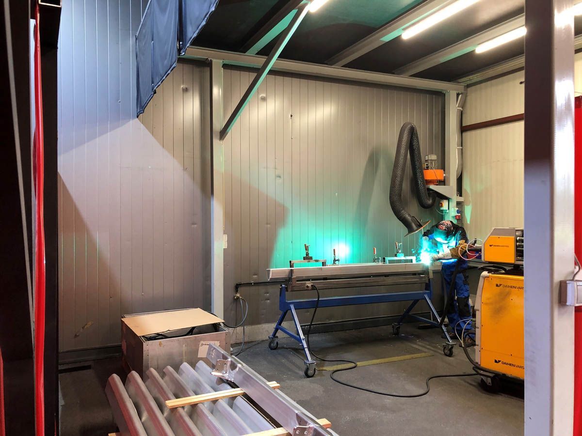 Schweißer bei der Arbeit in einer Werkstatt mit Metallwänden; grüne Schweißlampe, orange und blaue Ausrüstung.