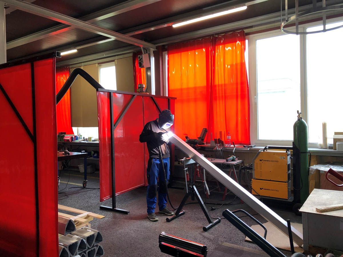 Eine Person schweißt in einer Werkstatt hinter einer roten Schutzwand einen langen Metallträger.