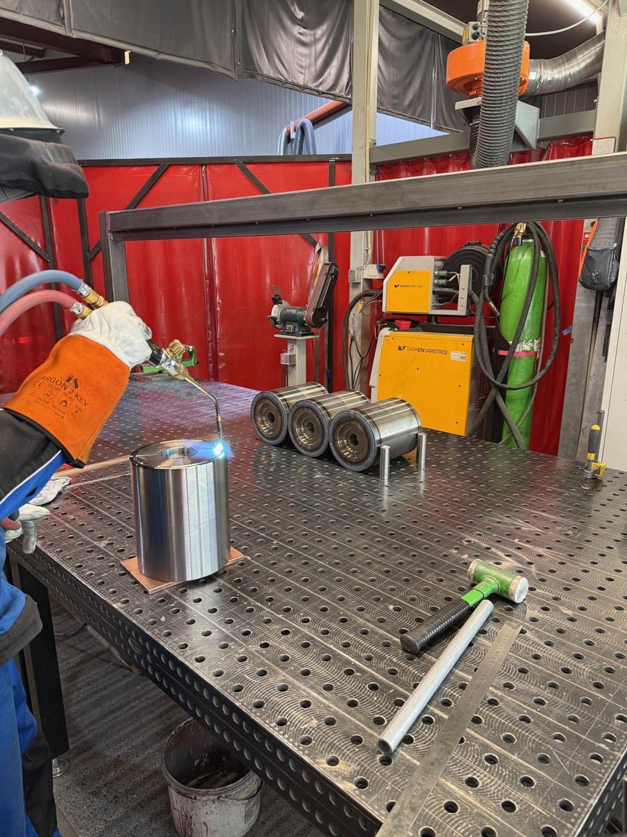 Ein Schweißer bearbeitet mit einem Schweißbrenner ein zylindrisches Metallobjekt auf einem Metalltisch in einer Werkstatt.