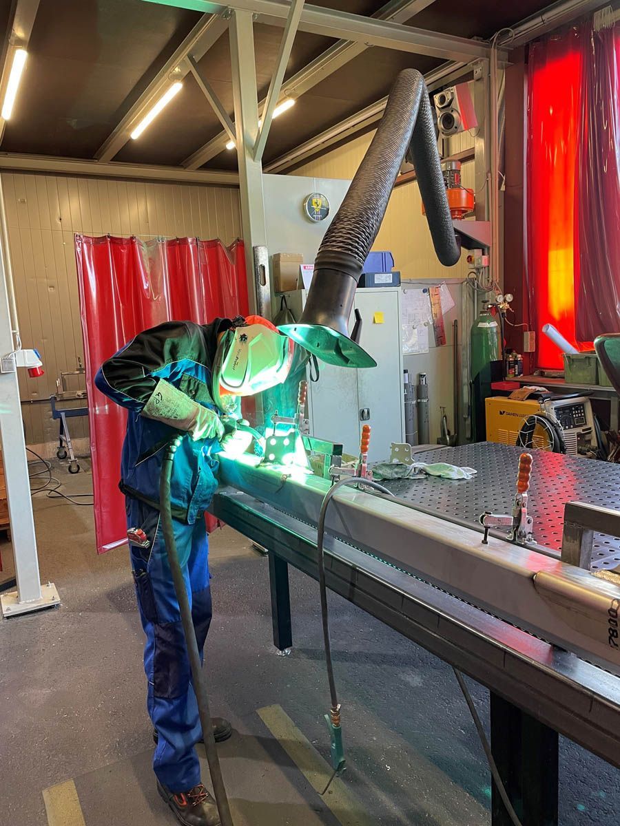 Schweißer in Schutzkleidung schweißt Metall in einer Werkstatt mit Belüftungssystem über der Decke.