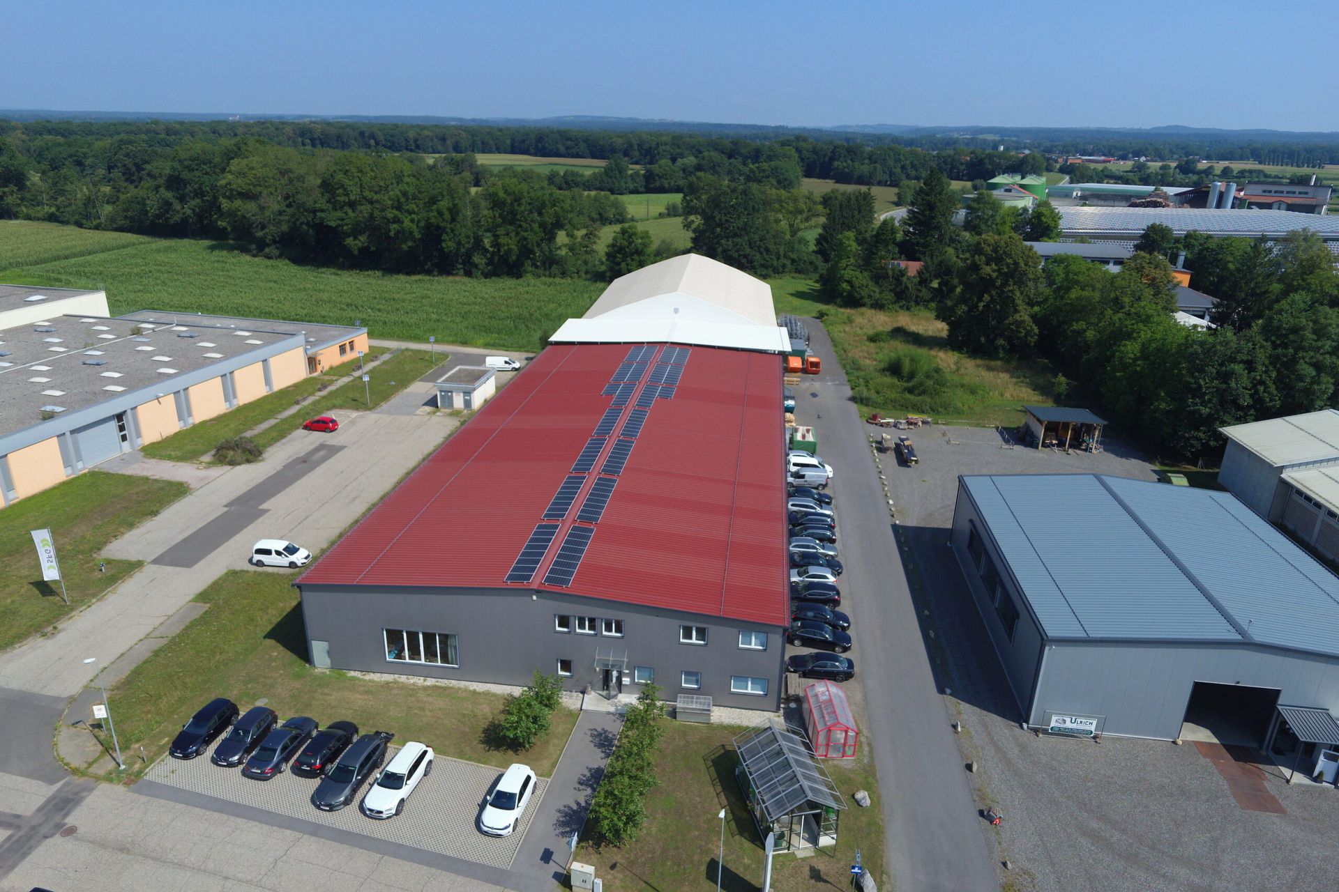 Luftaufnahme von Industriegebäuden mit roten und grauen Dächern, umgeben von Bäumen und einem Parkplatz.