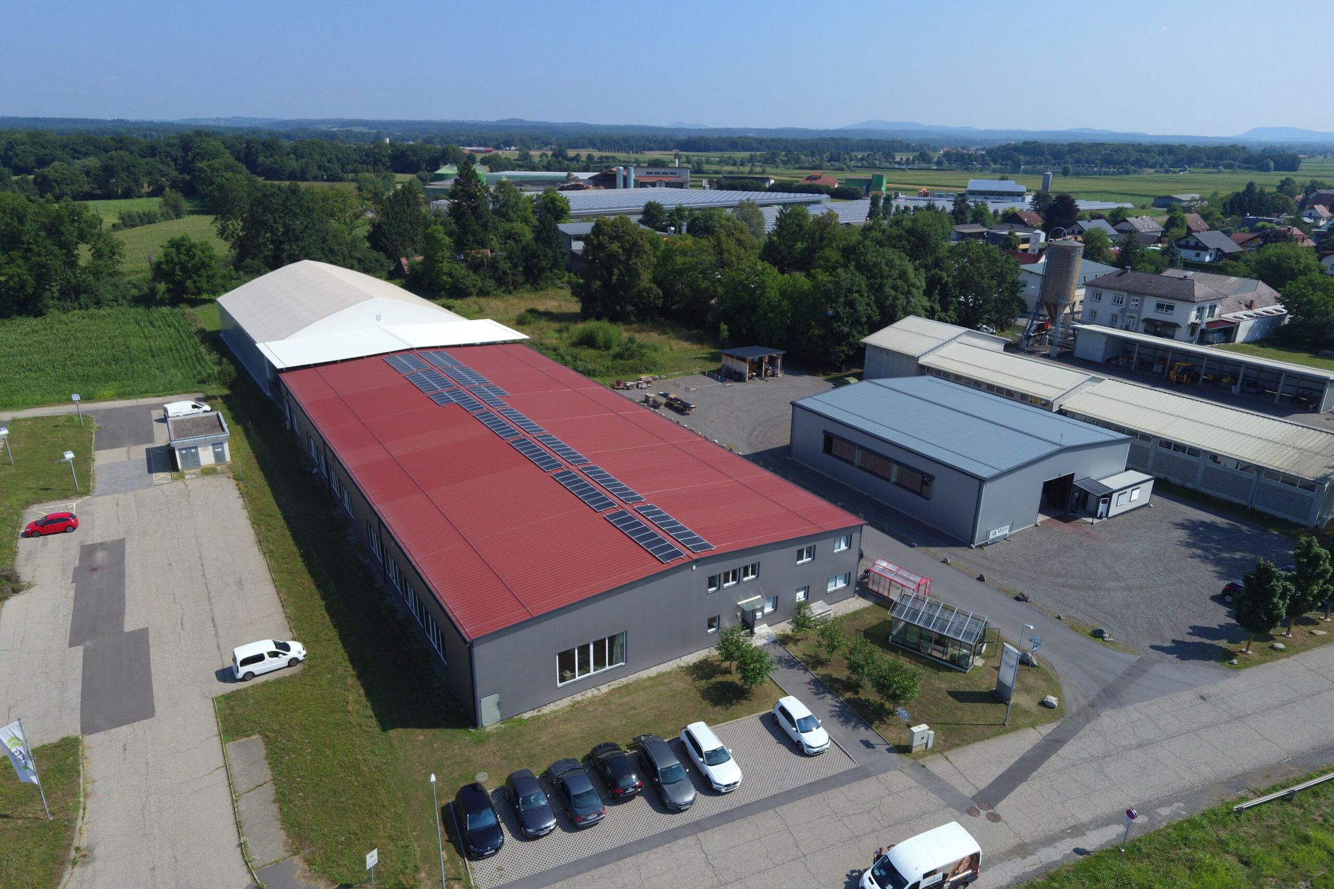 Luftaufnahme eines langen, rotgedeckten Industriegebäudes und kleinerer angrenzender Gebäude mit davor geparkten Autos.