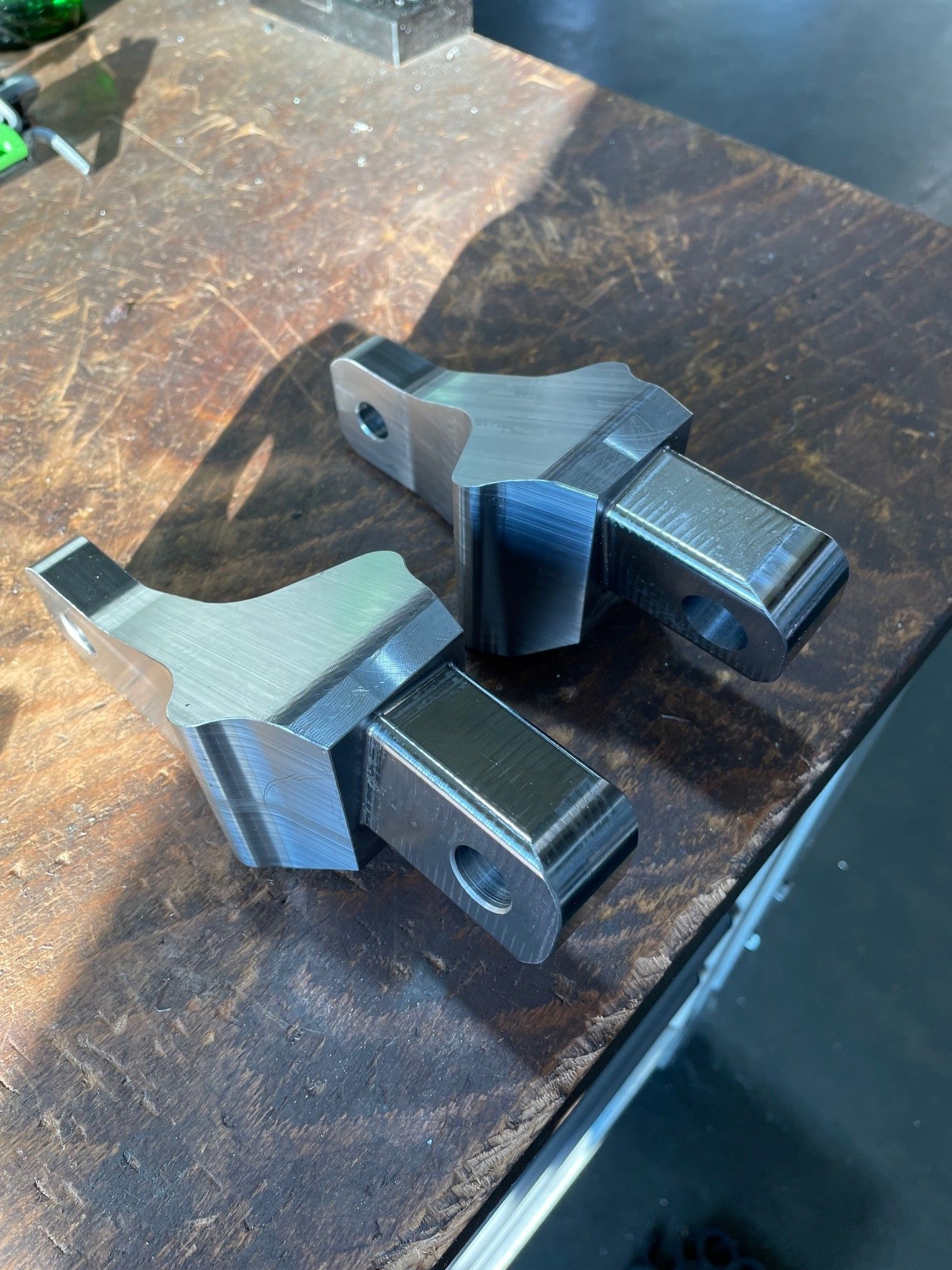 Zwei maschinell bearbeitete, Y-förmige Metallteile mit Löchern, die auf einer abgenutzten Holzoberfläche liegen.