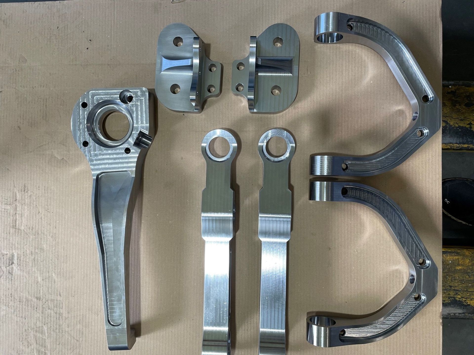 Bearbeitete Metallbauteile für die Fahrzeugaufhängung, darunter Arme, Halterungen und Stützen, auf hellbrauner Oberfläche.