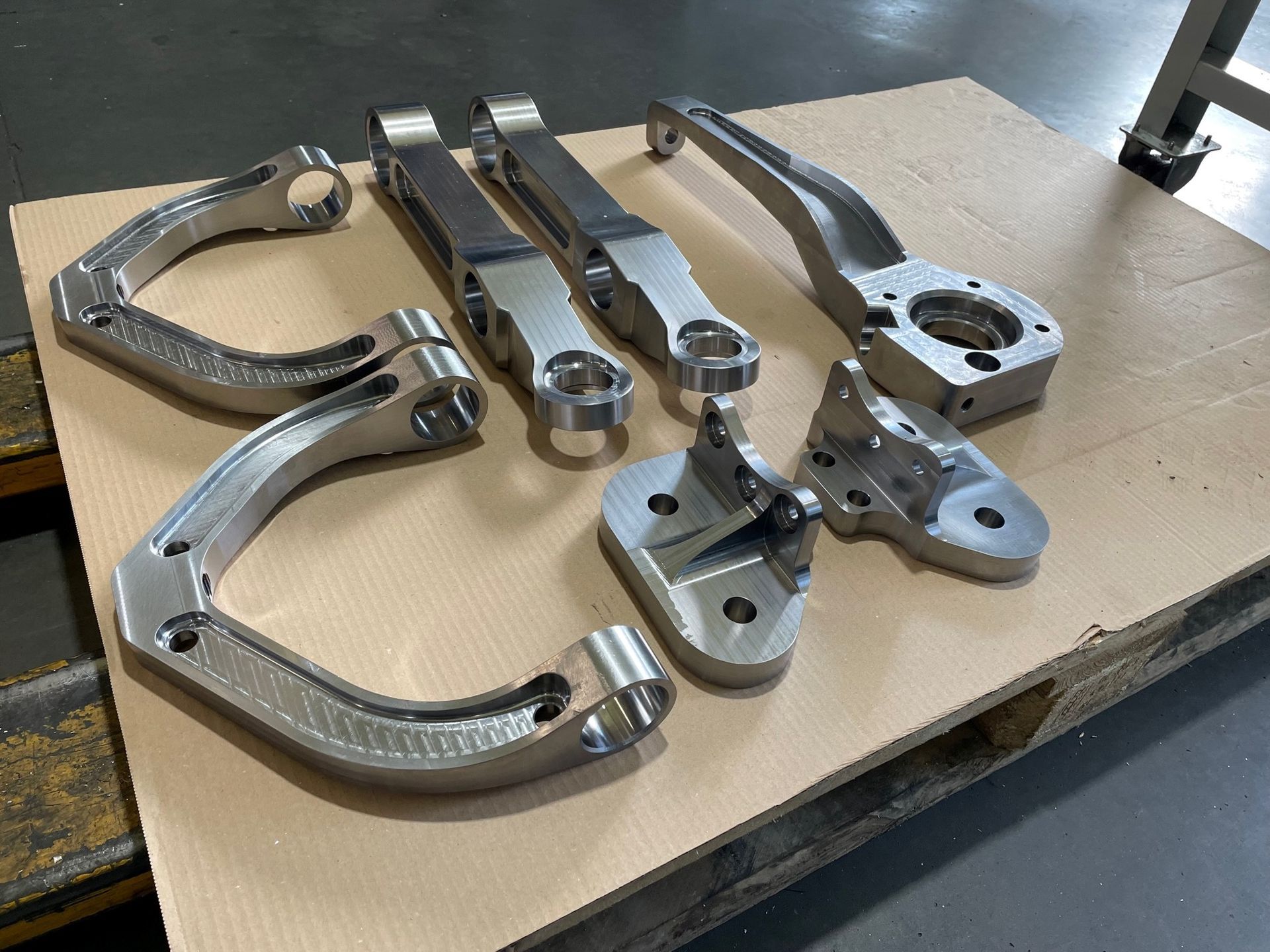 Mehrere bearbeitete Metallaufhängungskomponenten sind in einer Fabrikumgebung auf einer Holzpalette angeordnet.