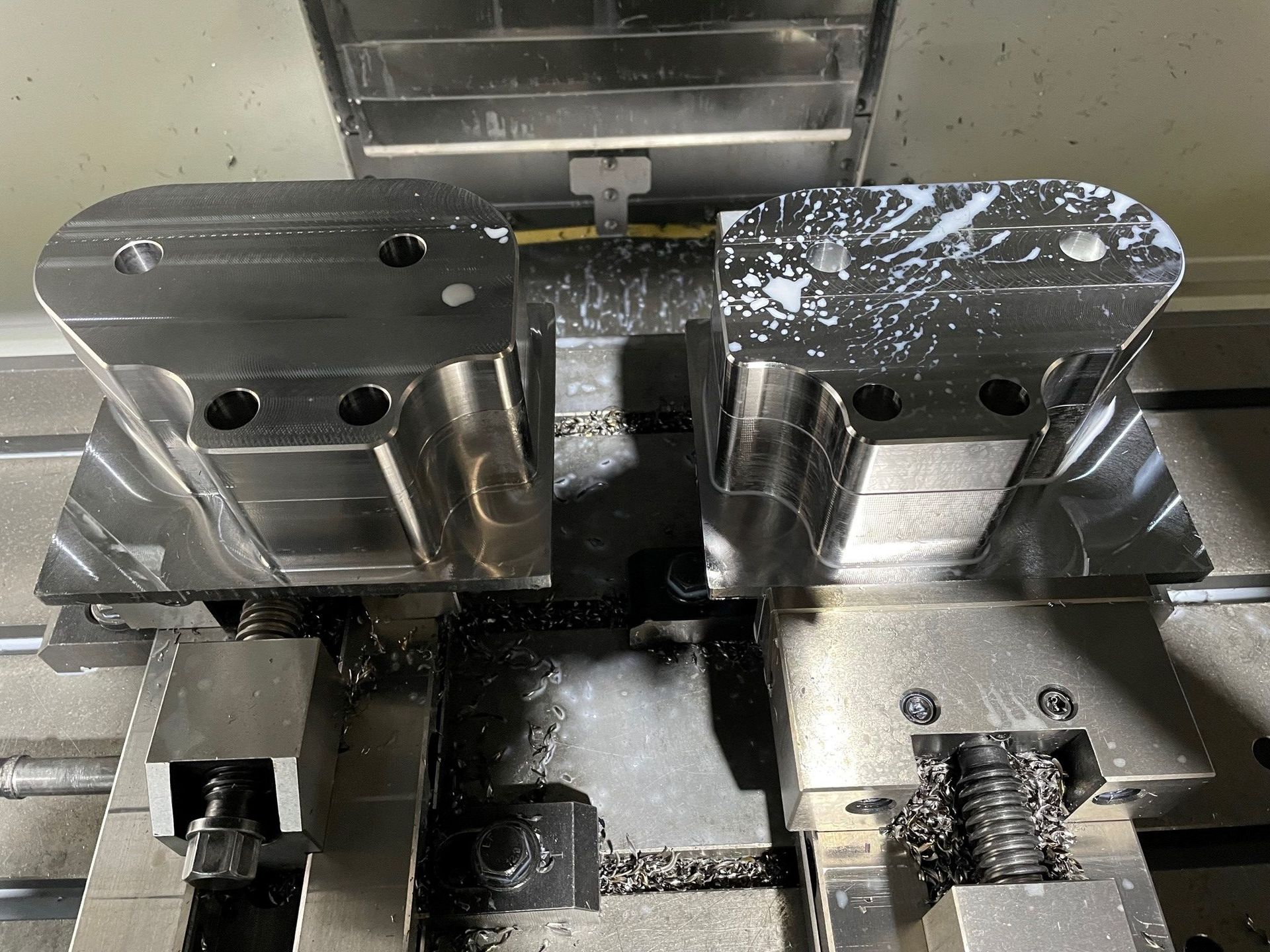 Zwei in einer CNC-Maschine eingespannte Metallteile, bedeckt mit Kühlschmierstoff und Metallspänen.