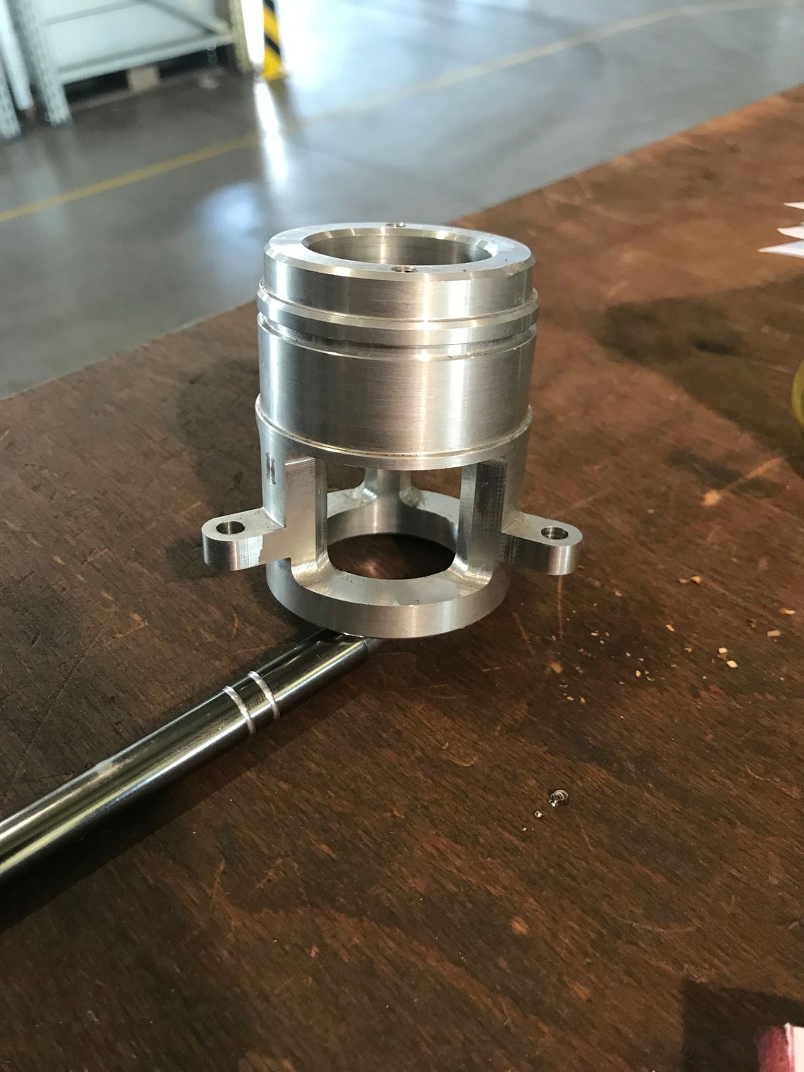 Ein silberfarbenes, maschinell bearbeitetes Metallteil liegt auf einem Holztisch; ein Stift zeigt auf den Sockel.