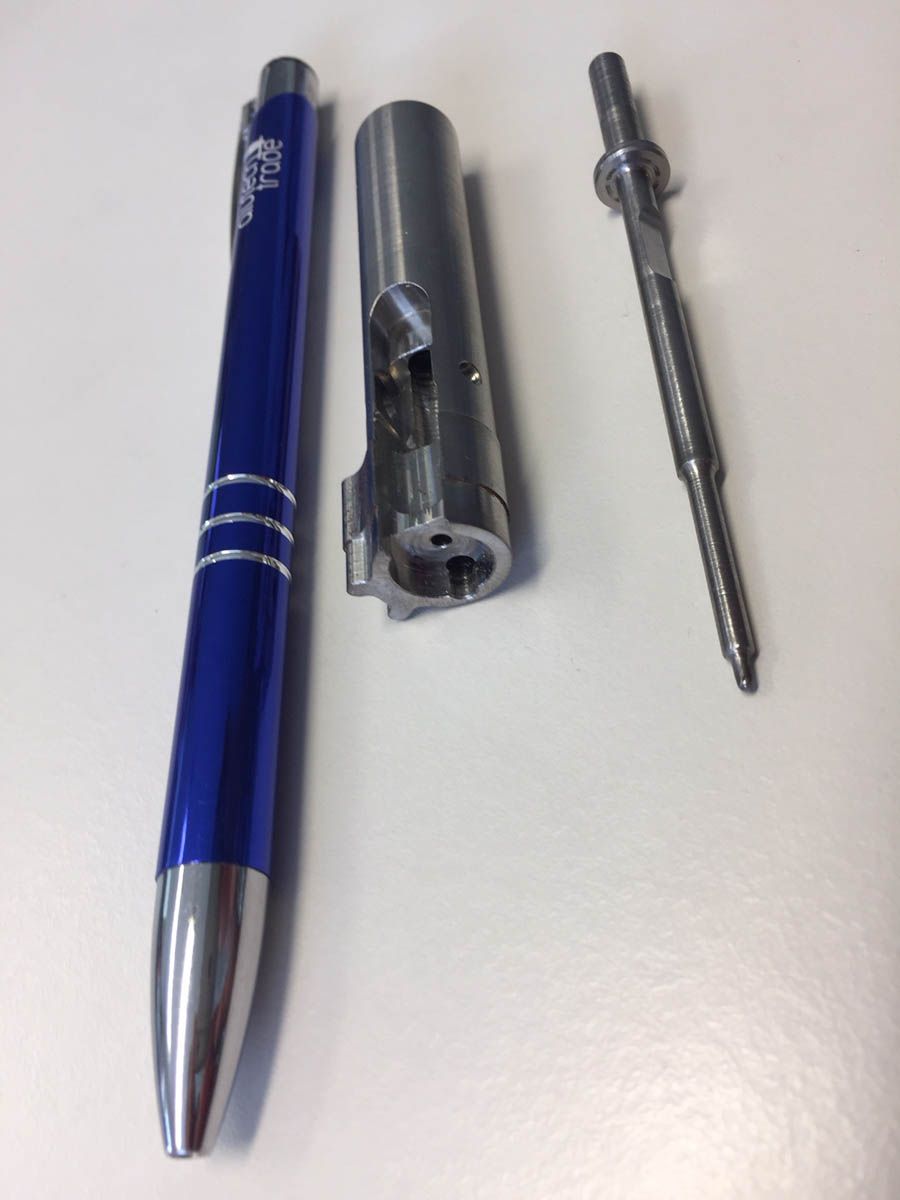 Drei metallene Schusswaffenteile, darunter ein Verschluss und ein Schlagbolzen, mit einem blauen Stift auf weißem Untergrund.