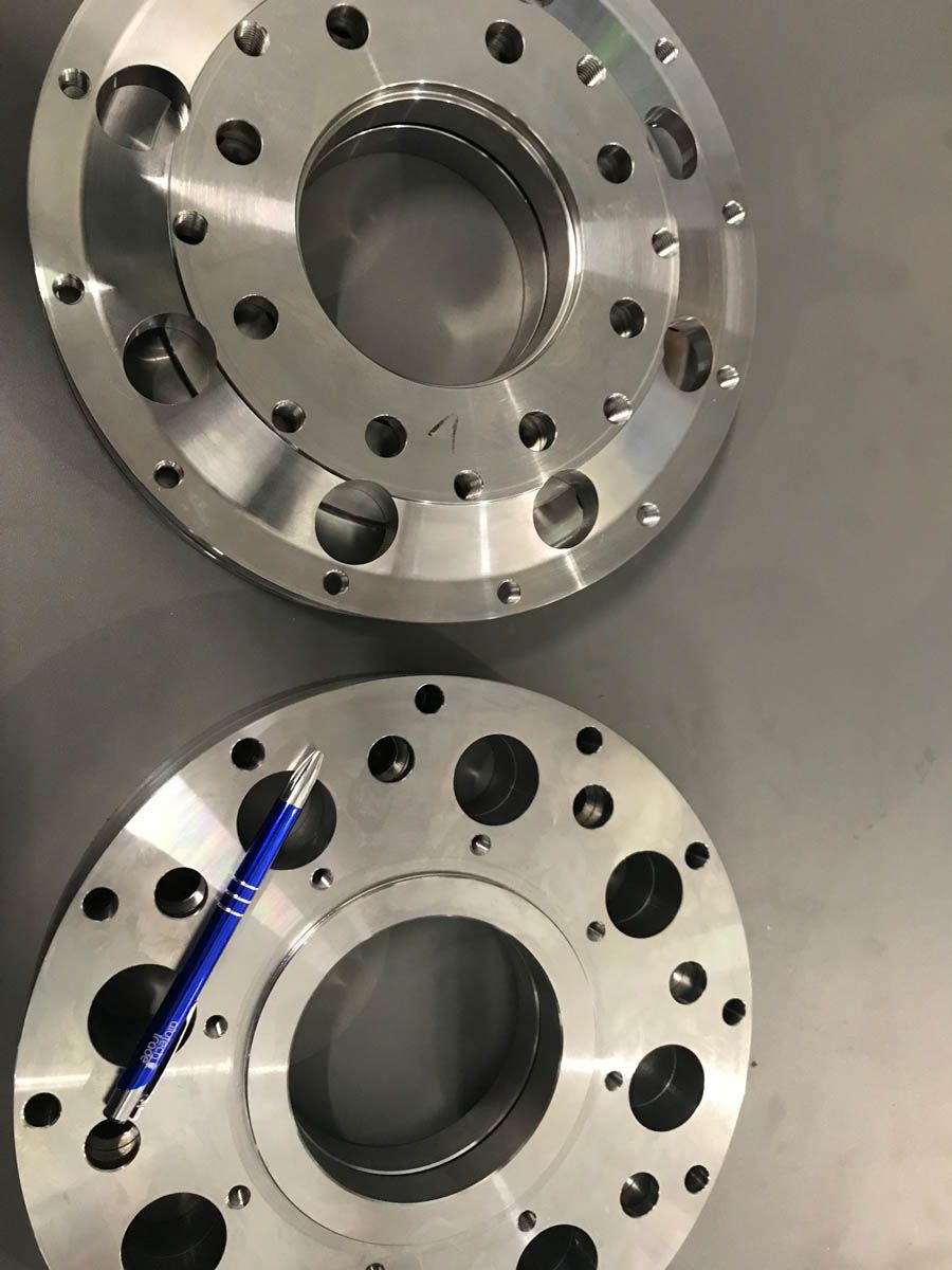 Zwei bearbeitete Metallflanschplatten mit verschiedenen Löchern und runden Öffnungen. Auf einer davon liegt ein Stift.