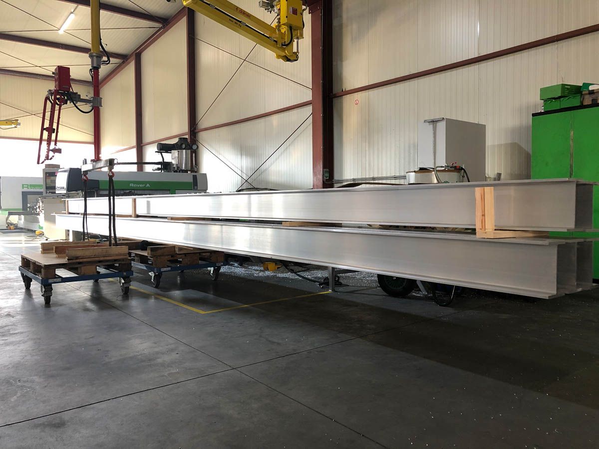 Große Metallträger sind in einer Fabrikumgebung, möglicherweise für Bauzwecke, in der Nähe einer Schneidemaschine gestapelt.