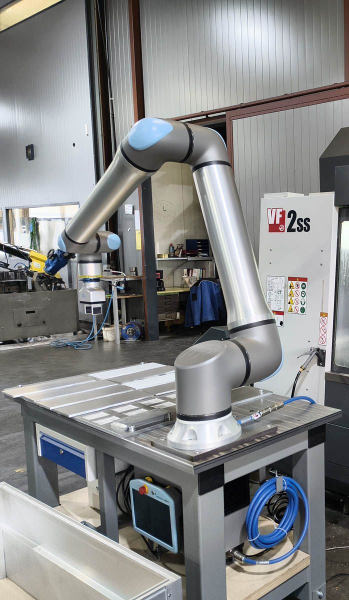 Roboterarm in einer Werkstattumgebung, grauer Arm, der sich über einen Metalltisch mit Bedienfeld und Werkzeugen erstreckt.