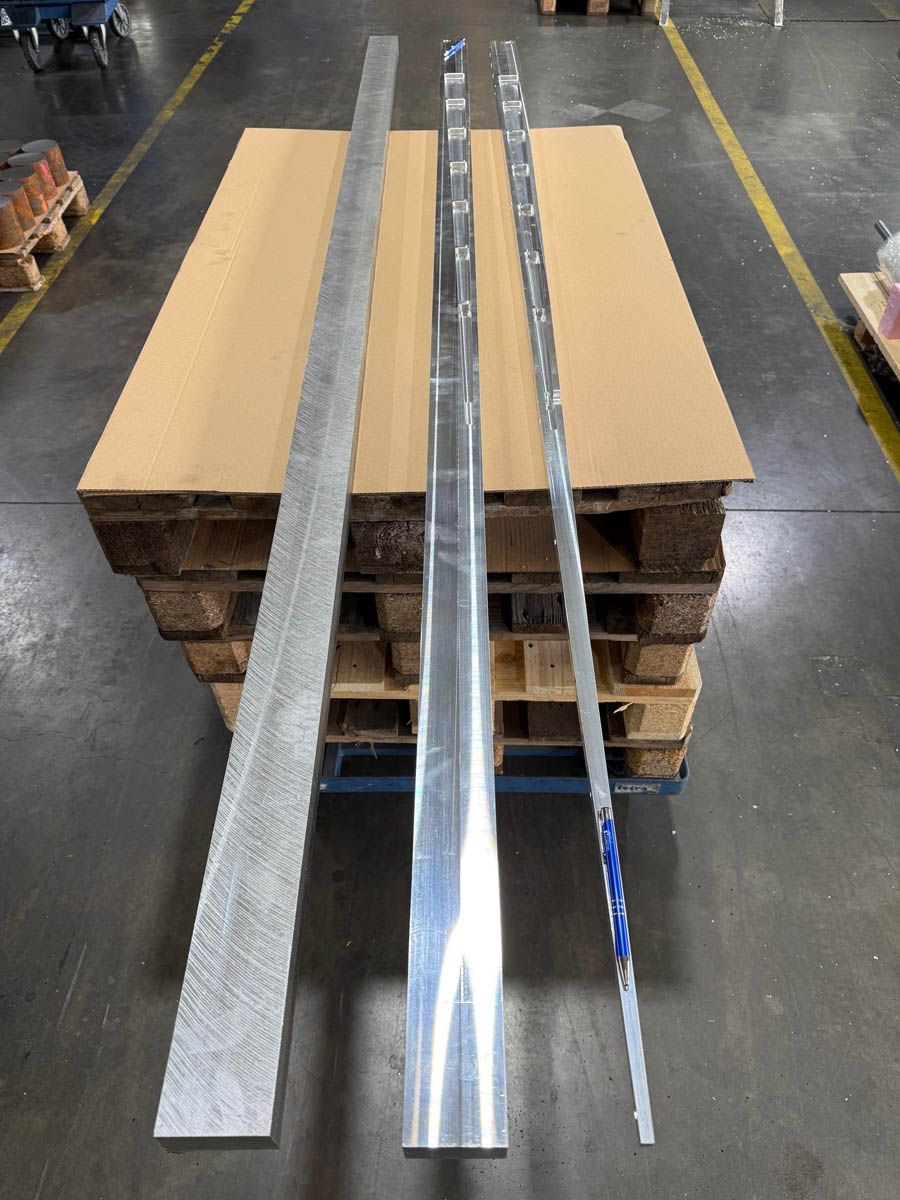 Drei lange Metallträger unterschiedlicher Bauart liegen in einer Fabrik auf einer Holzpalette.