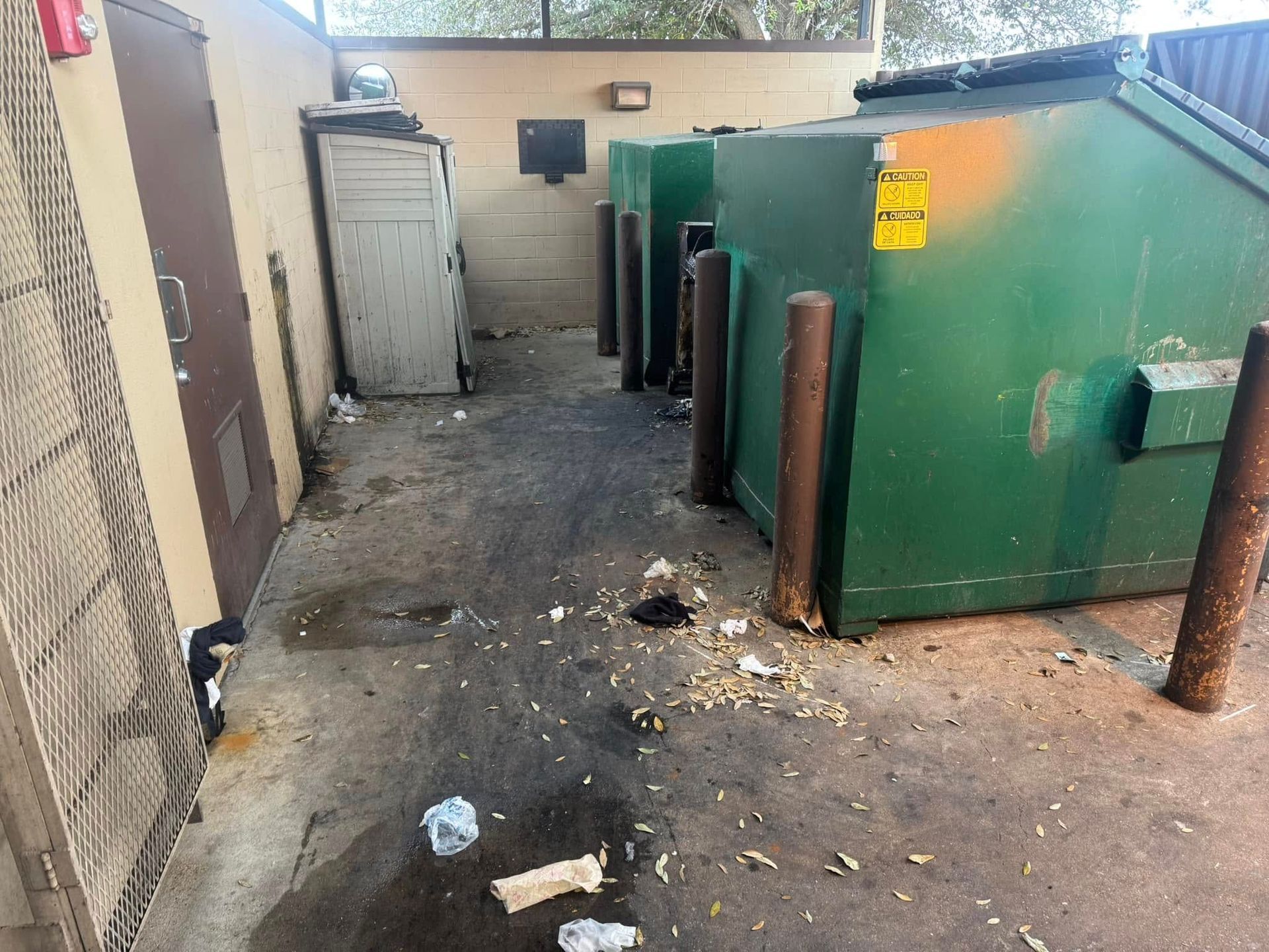 A narrow outdoor area with two green industrial dumpsters, a utility door, and loose trash scattered on the ground.