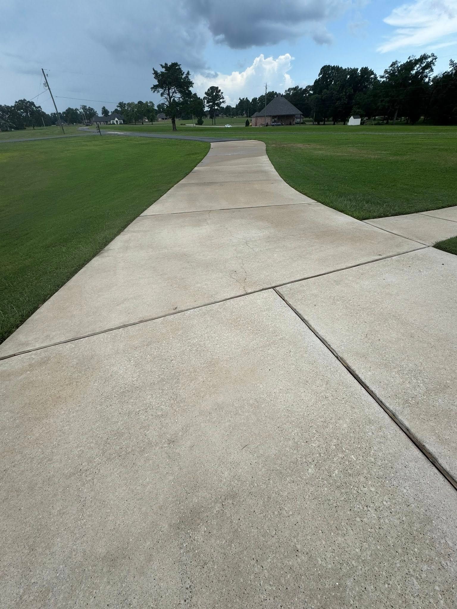 A concrete sidewalk curves through a grassy field toward a distant building under a cloudy, bright blue sky.