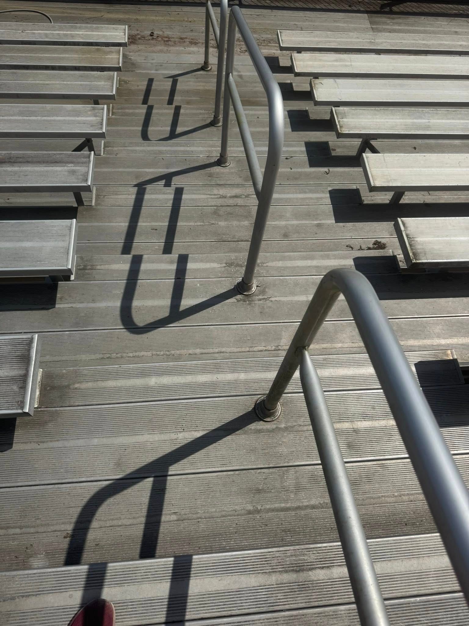 Metal bleachers with railings cast long, dark shadows across the wooden seating area on a sunny day.