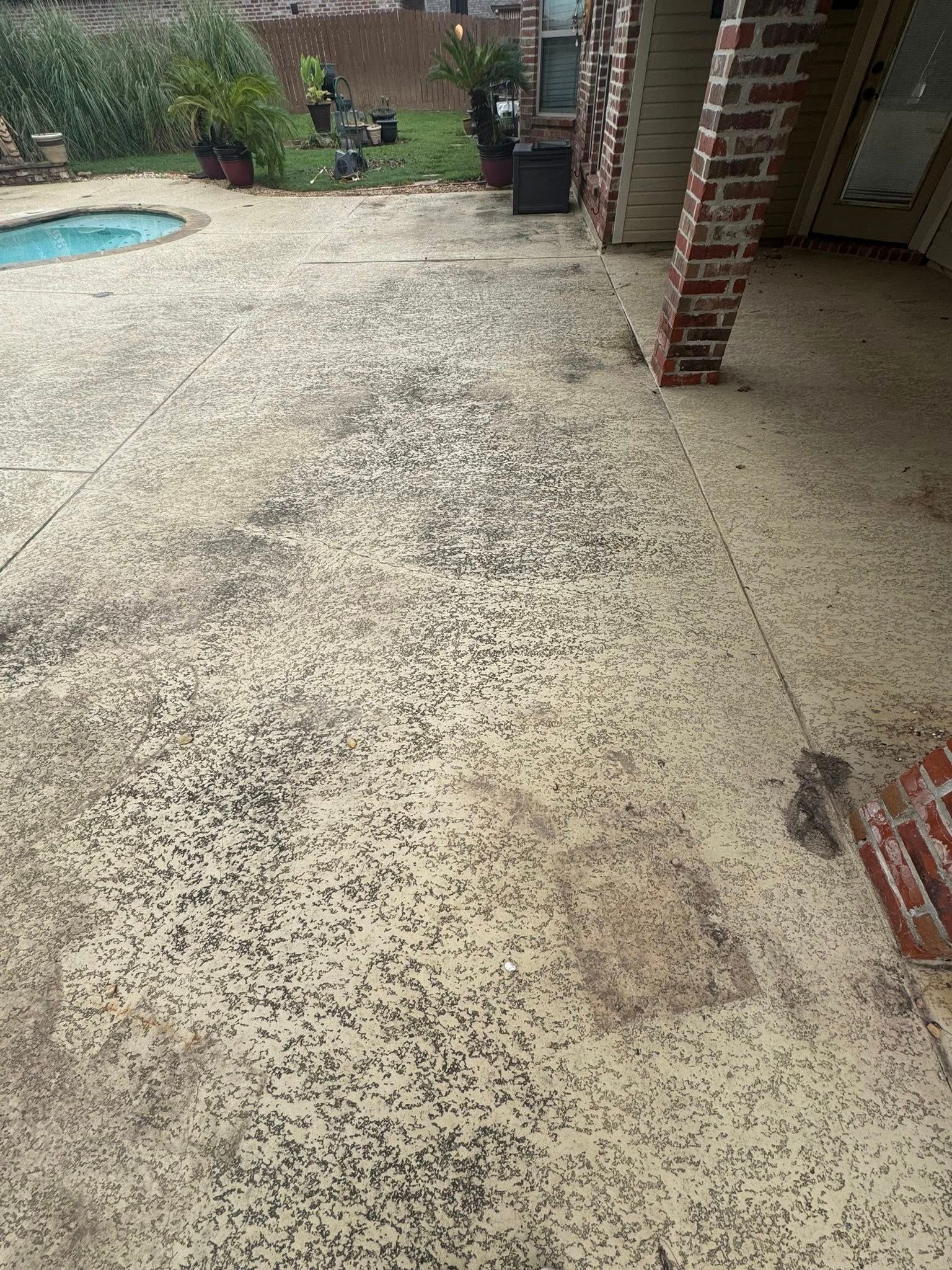 A tan concrete patio with dark aggregate texture, leading toward a swimming pool, lawn, and a brick-pillared patio cover.