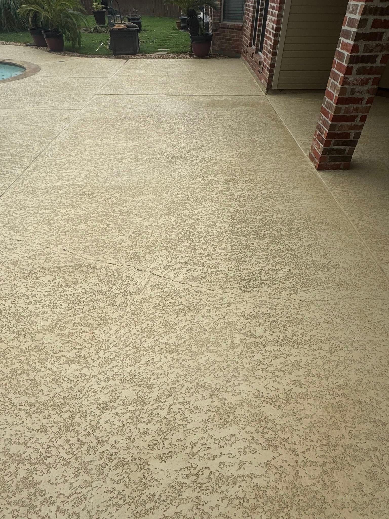 A close-up view of a textured, light-colored concrete patio surface with brown decorative flecks next to a brick pillar.