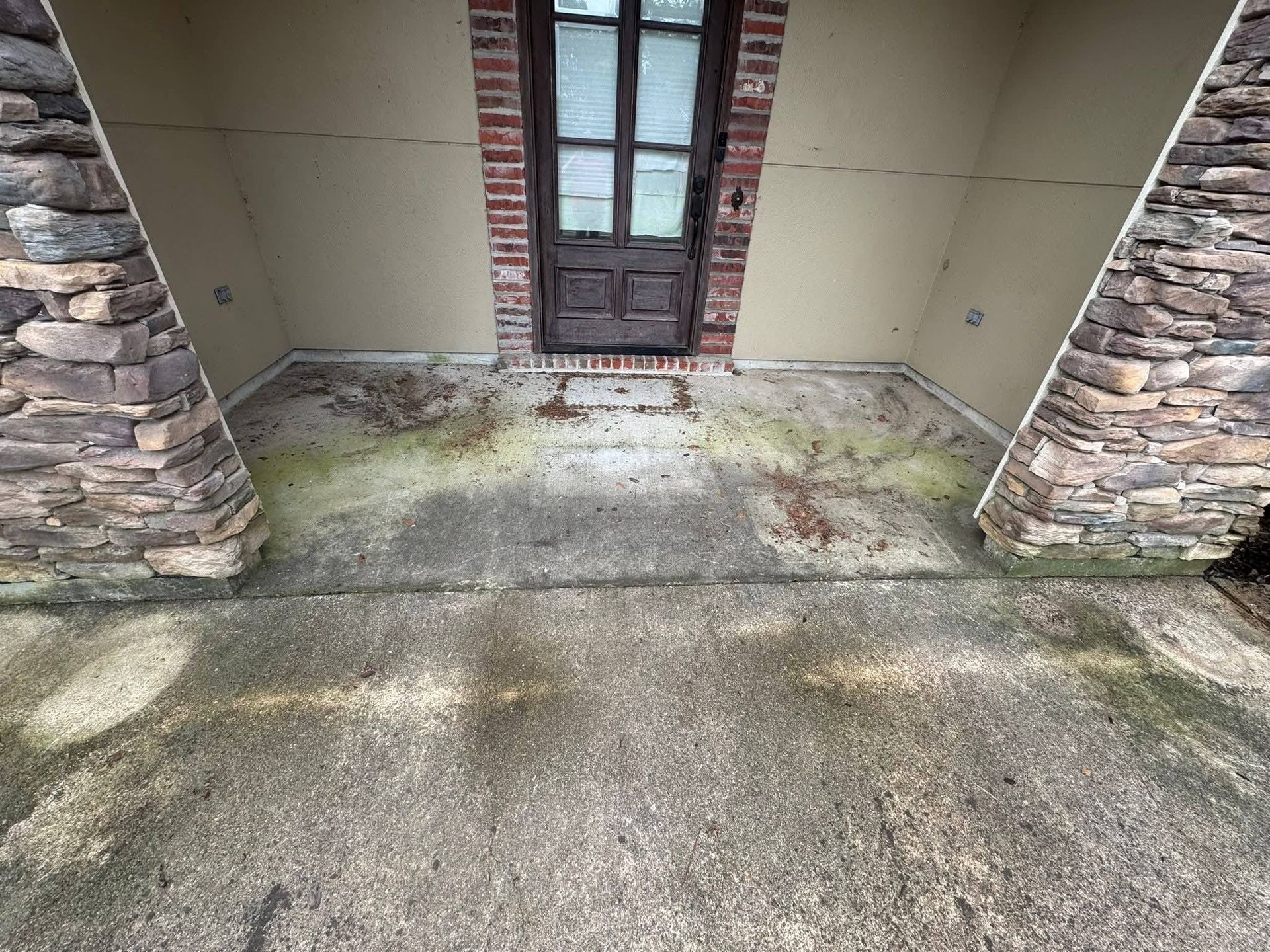 An entryway with a dark wooden door framed in brick, flanked by stone columns, with a stained, worn concrete porch.