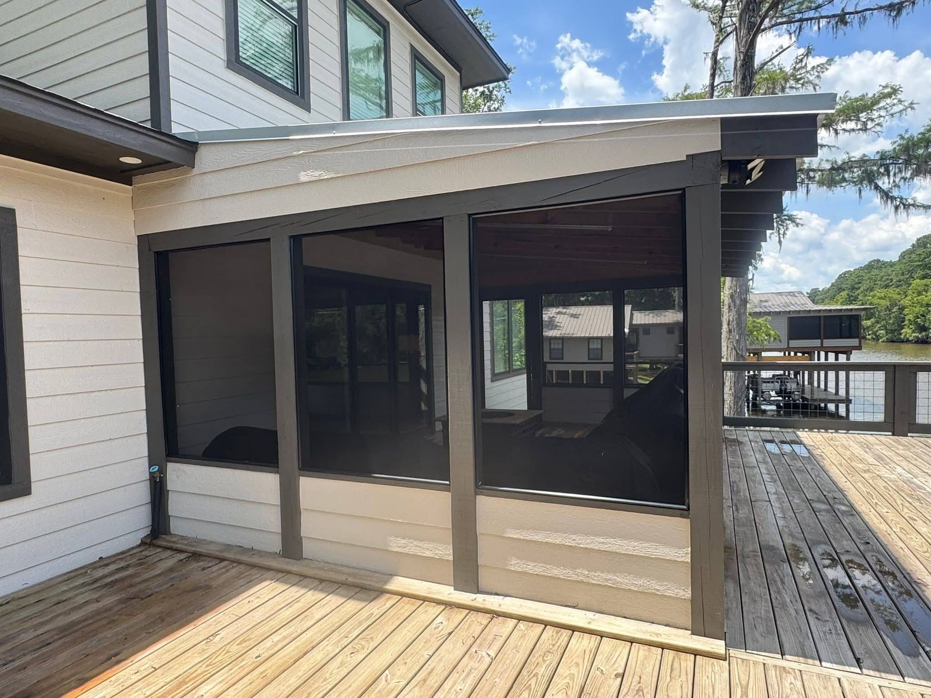 A screened-in porch with dark grey framing attached to a white, horizontal-sided house overlooking a lake with a wooden deck.