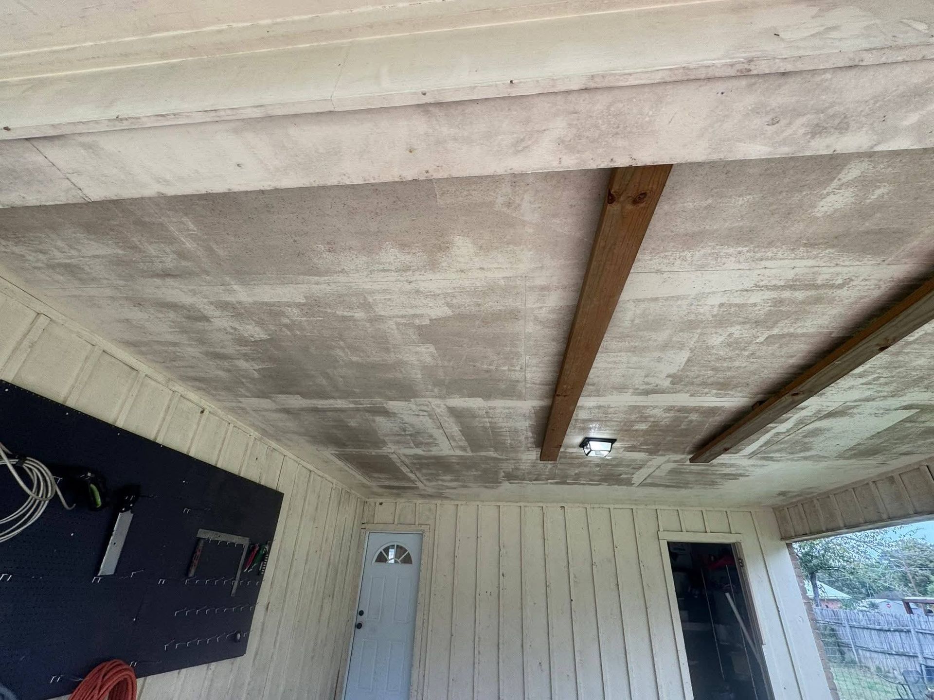 A view of a porch ceiling made of textured panels divided by wood beams, with light-colored vertical walls and two doorways.