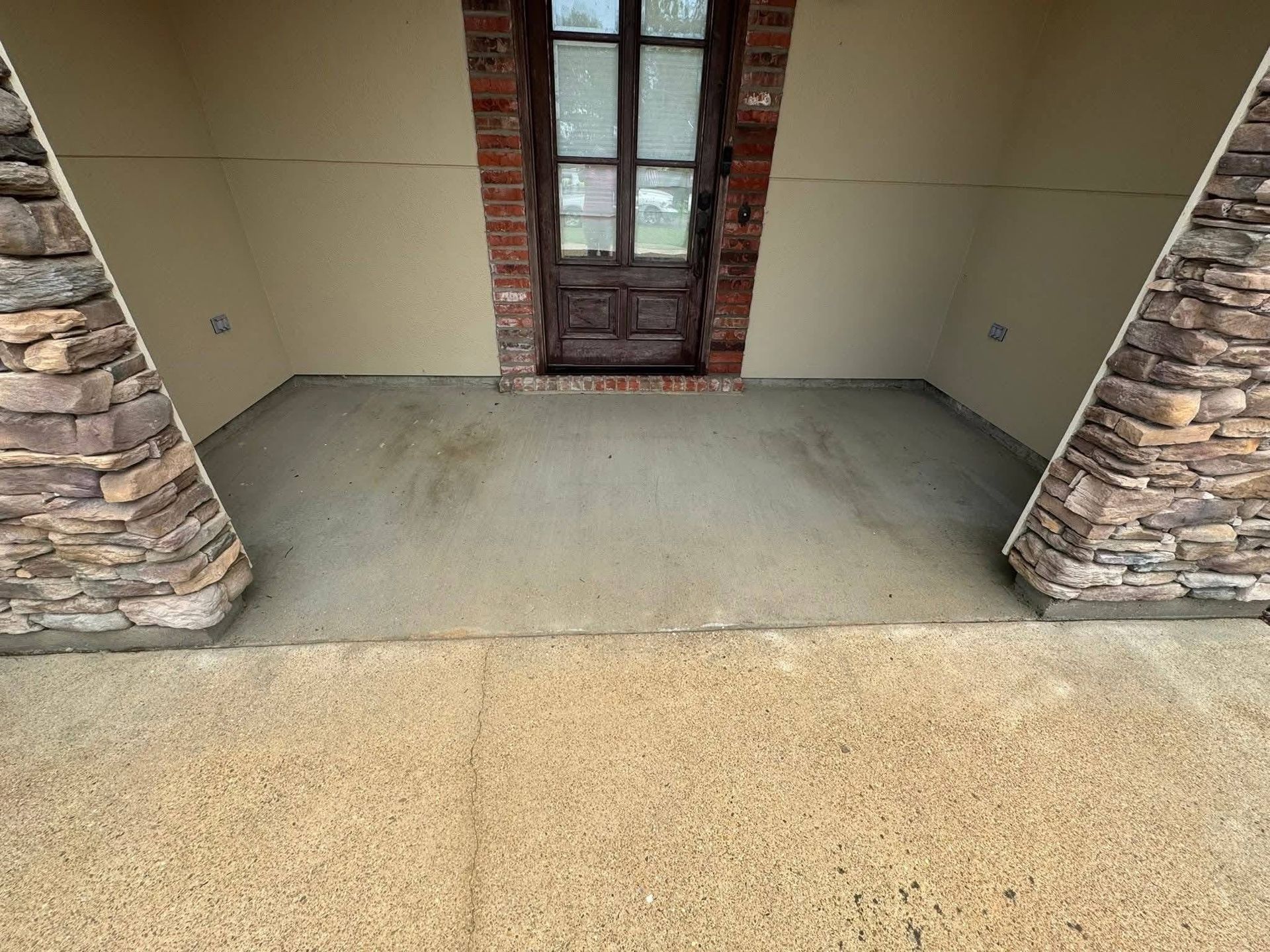 A covered front porch with a smooth concrete floor, flanked by stone columns, leading to a dark wood front door.
