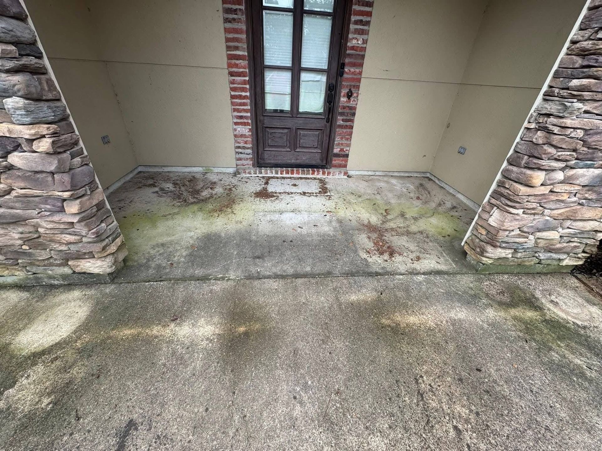 A front porch with a weathered concrete floor, stone pillars, and a brown wooden door framed by red brick.