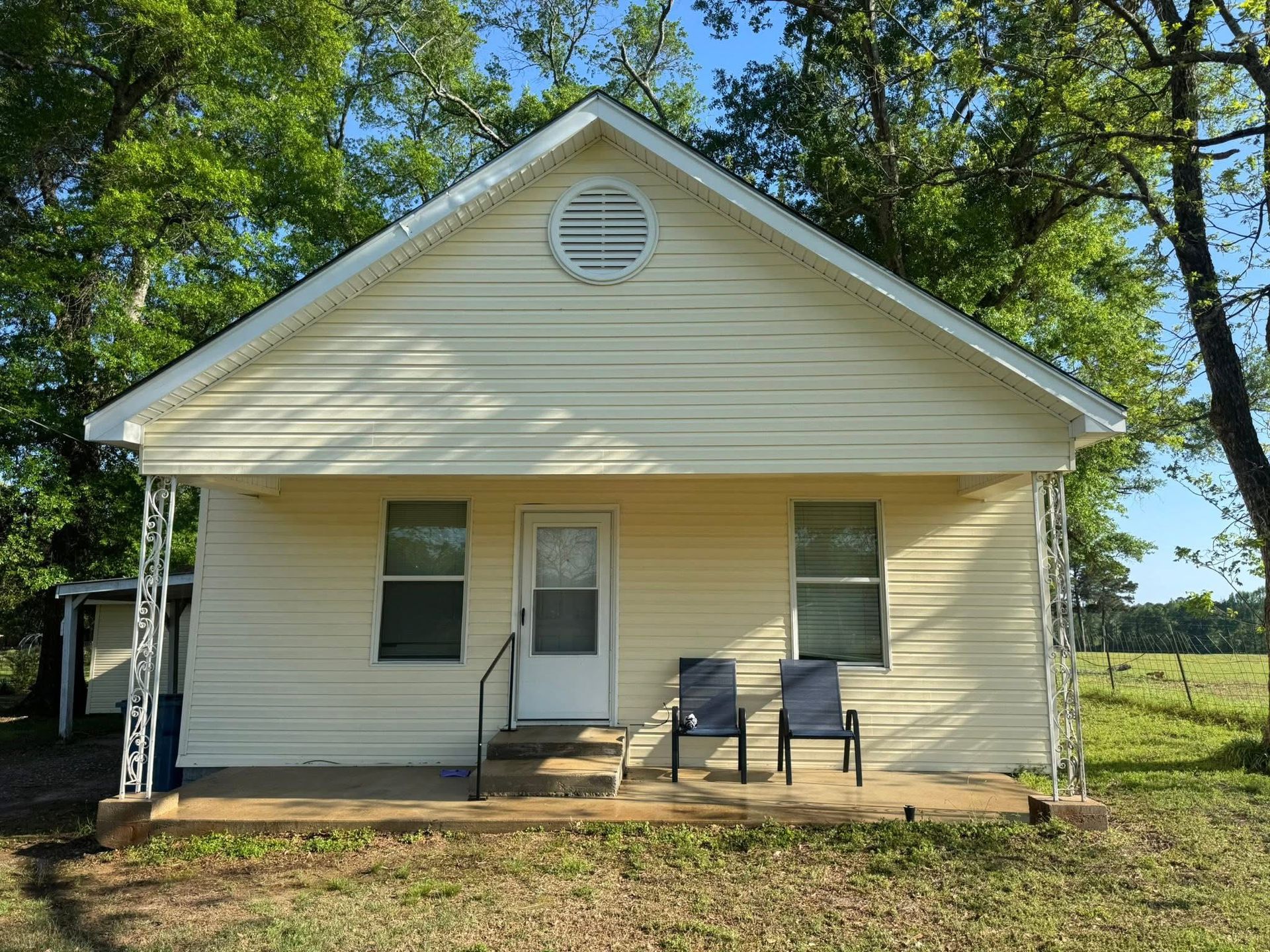 Small beige house with a front porch, two chairs, and trees in the yard.