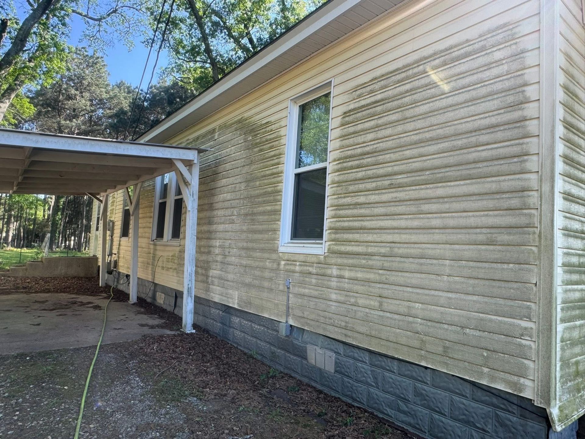 A side view of a light-yellow house with a carport, featuring weathered siding and a dark stone foundation.