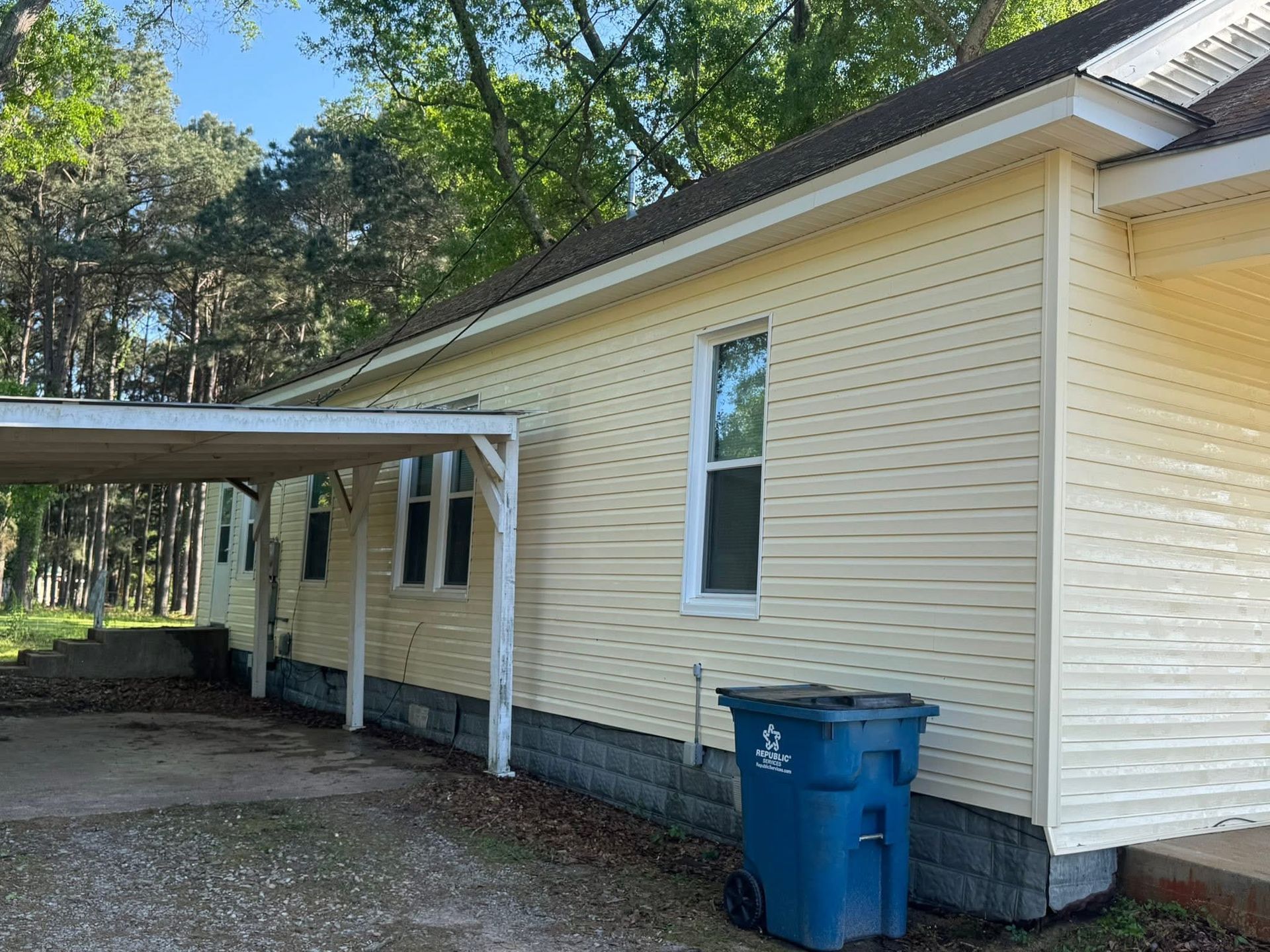 A side view of a light yellow house with white trim and a carport, with a blue trash bin against the gray brick foundation.