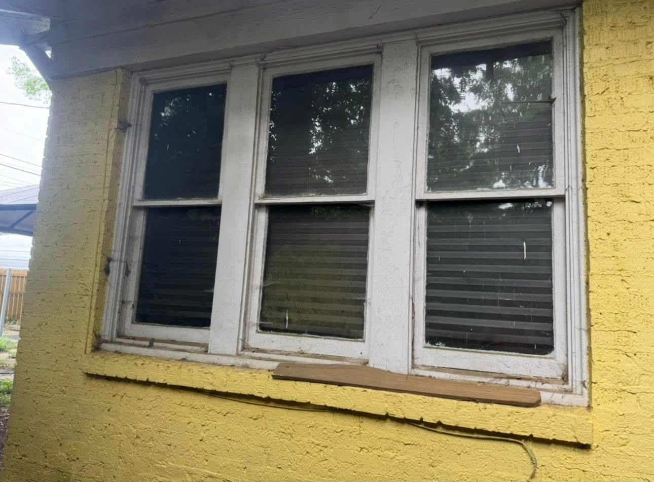 Three white-framed windows on a textured yellow stucco exterior wall, with a small piece of wood resting on the windowsill.