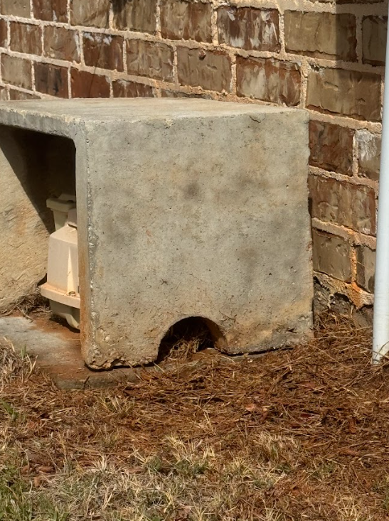 A concrete protective cover sitting on the ground against a brick wall, shielding a piece of utility equipment.