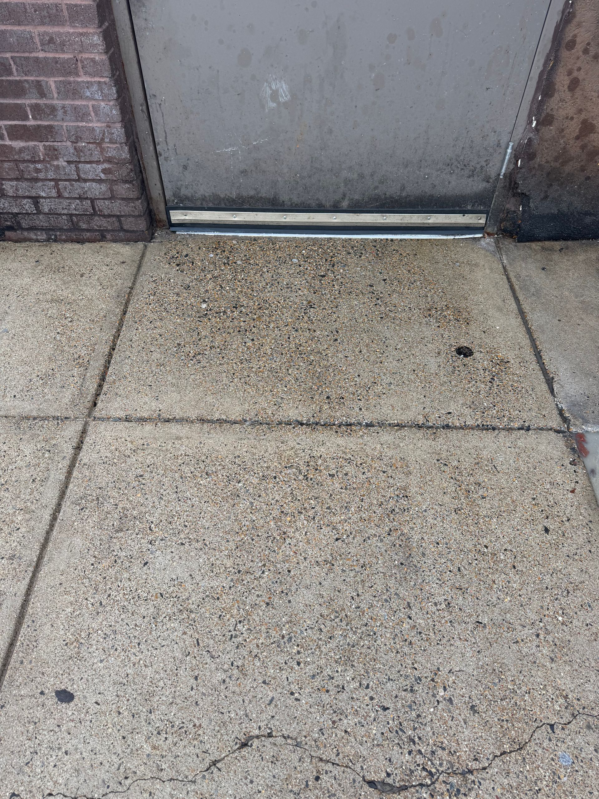 Concrete sidewalk leading to a gray metal door with a brick wall on the left.