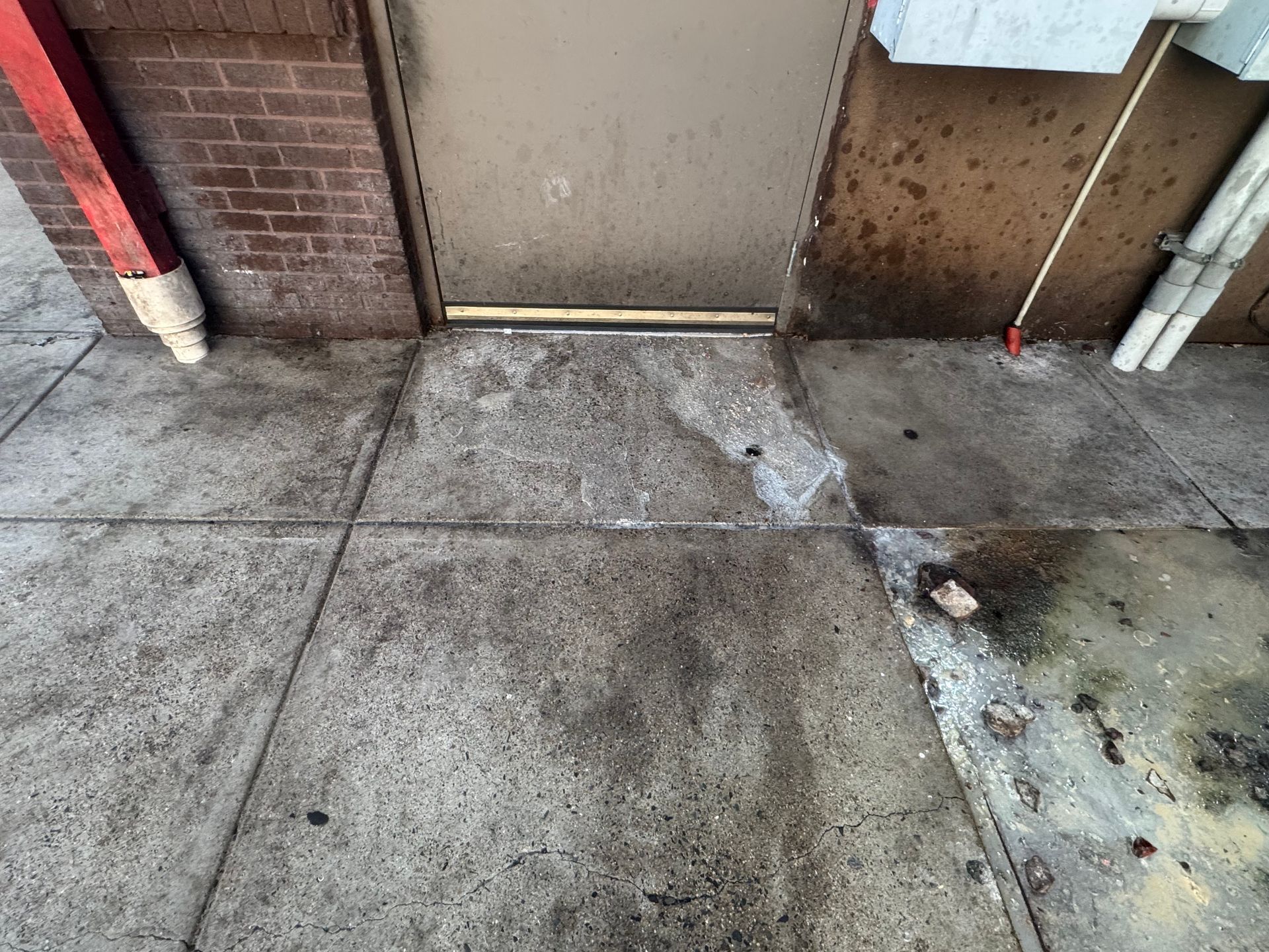 Concrete sidewalk beside a brick wall and door threshold, with cracks, stains, and debris.