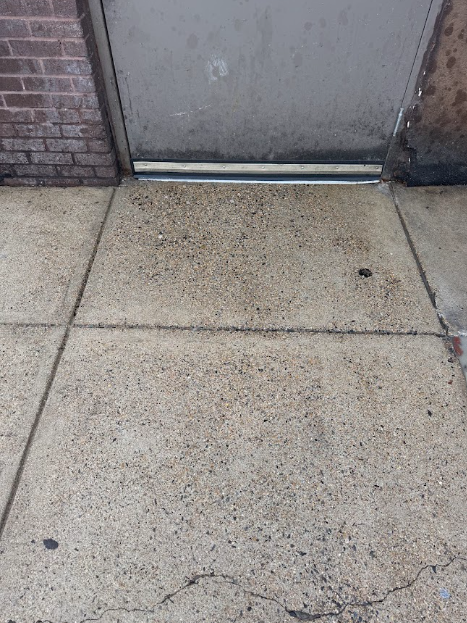 A concrete area between a beige wall and large green dumpsters, showing significant dark staining and wet patches on ground.