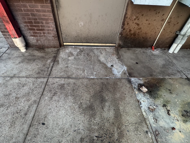A narrow outdoor area with two green industrial dumpsters, a utility door, and loose trash scattered on the ground.