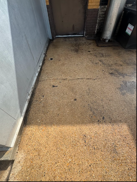 A concrete area between a beige wall and large green dumpsters, showing significant dark staining and wet patches on ground.