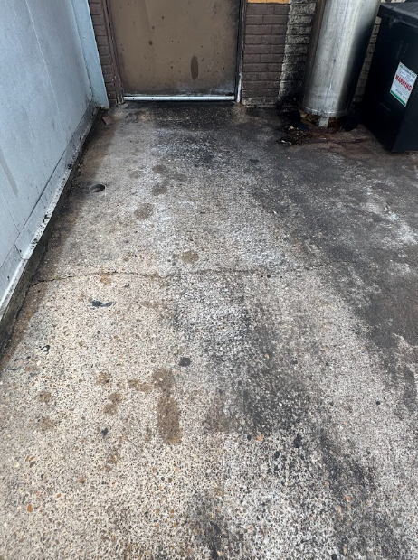 A narrow outdoor area with two green industrial dumpsters, a utility door, and loose trash scattered on the ground.