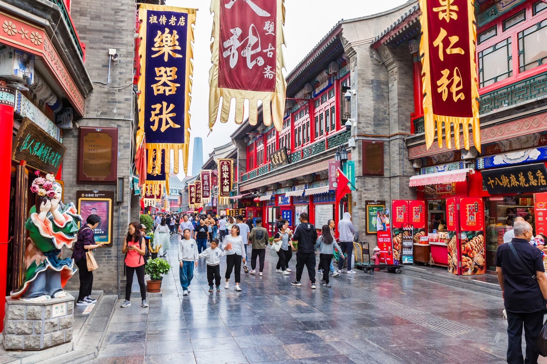 Calle en China con edificios rojos, gente caminando y pancartas colgando en lo alto.