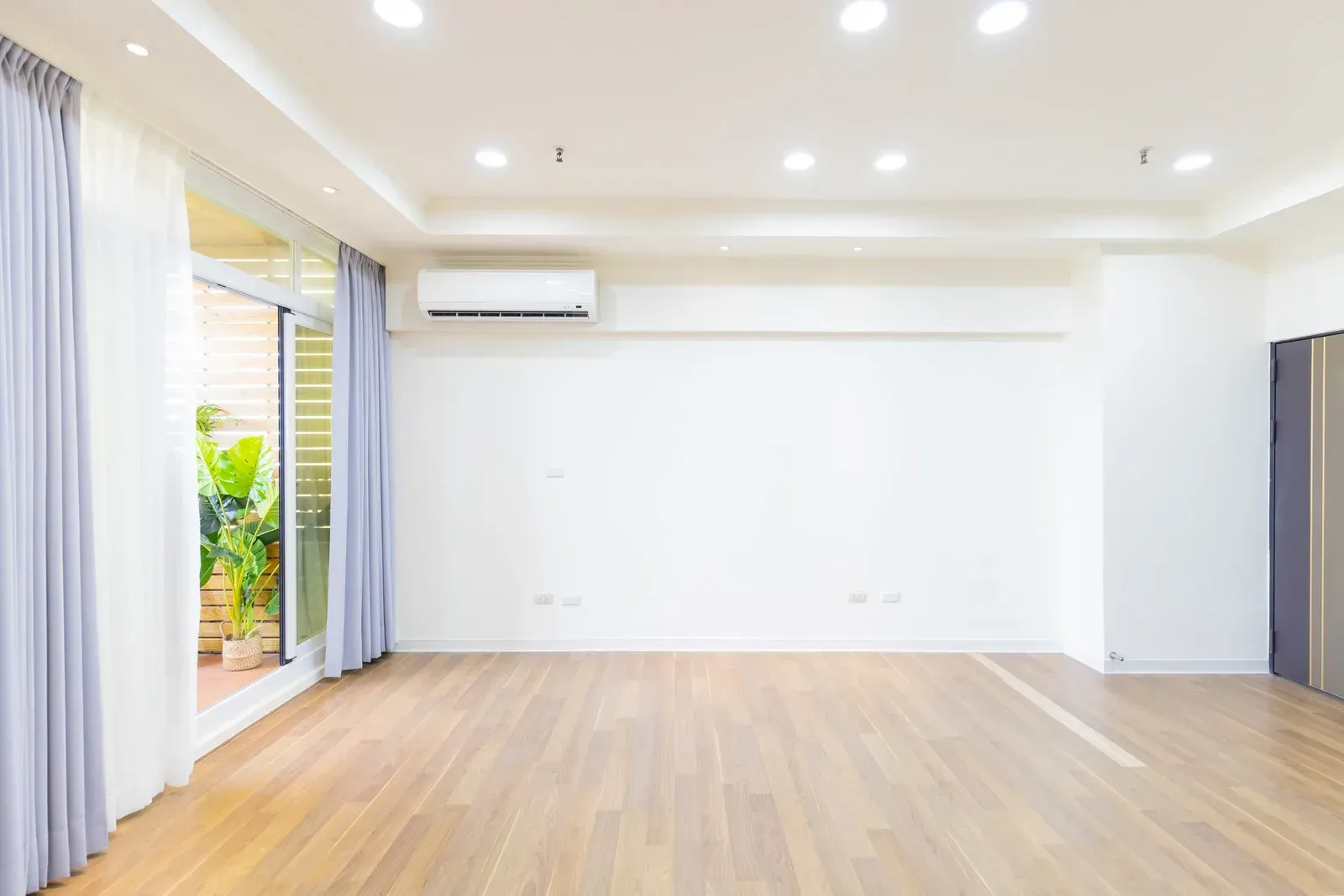 Empty, bright room with hardwood floor, sliding glass door, white walls, AC unit, and gray curtains.