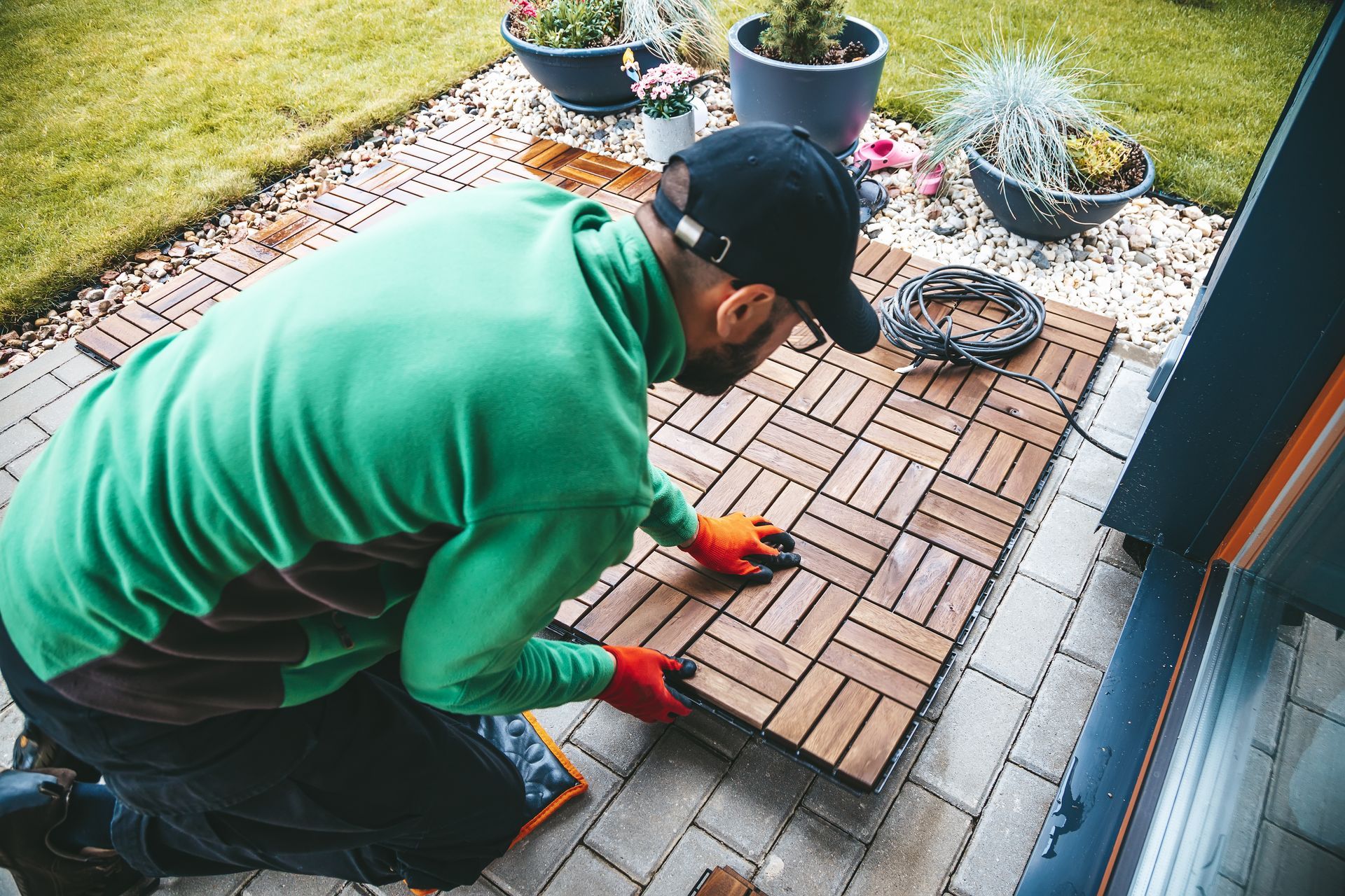 Person in green hoodie laying wooden pavers on a patio beside grass and potted plants