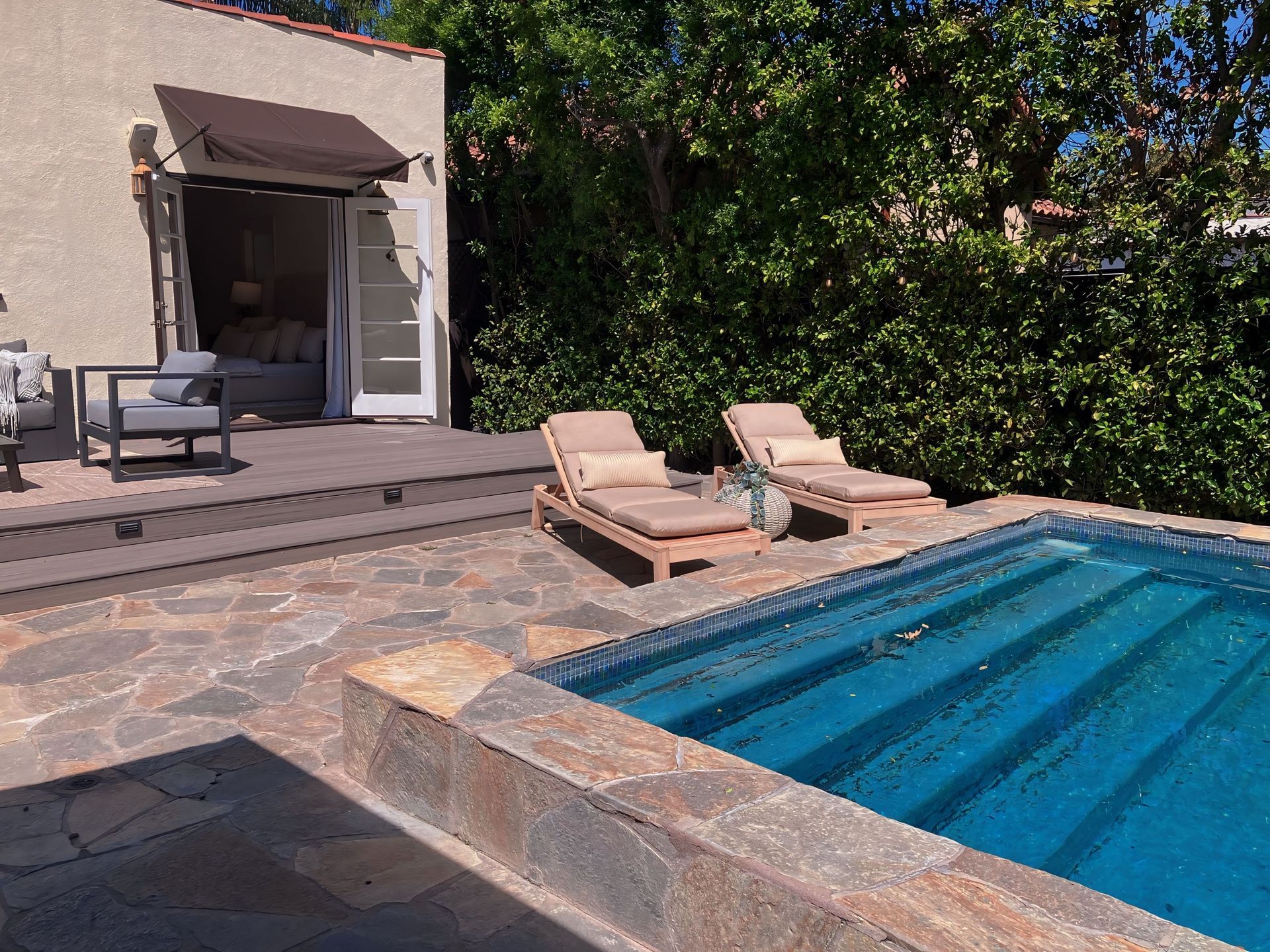 Backyard pool with two lounge chairs, stone patio, and a covered seating area by a hedge
