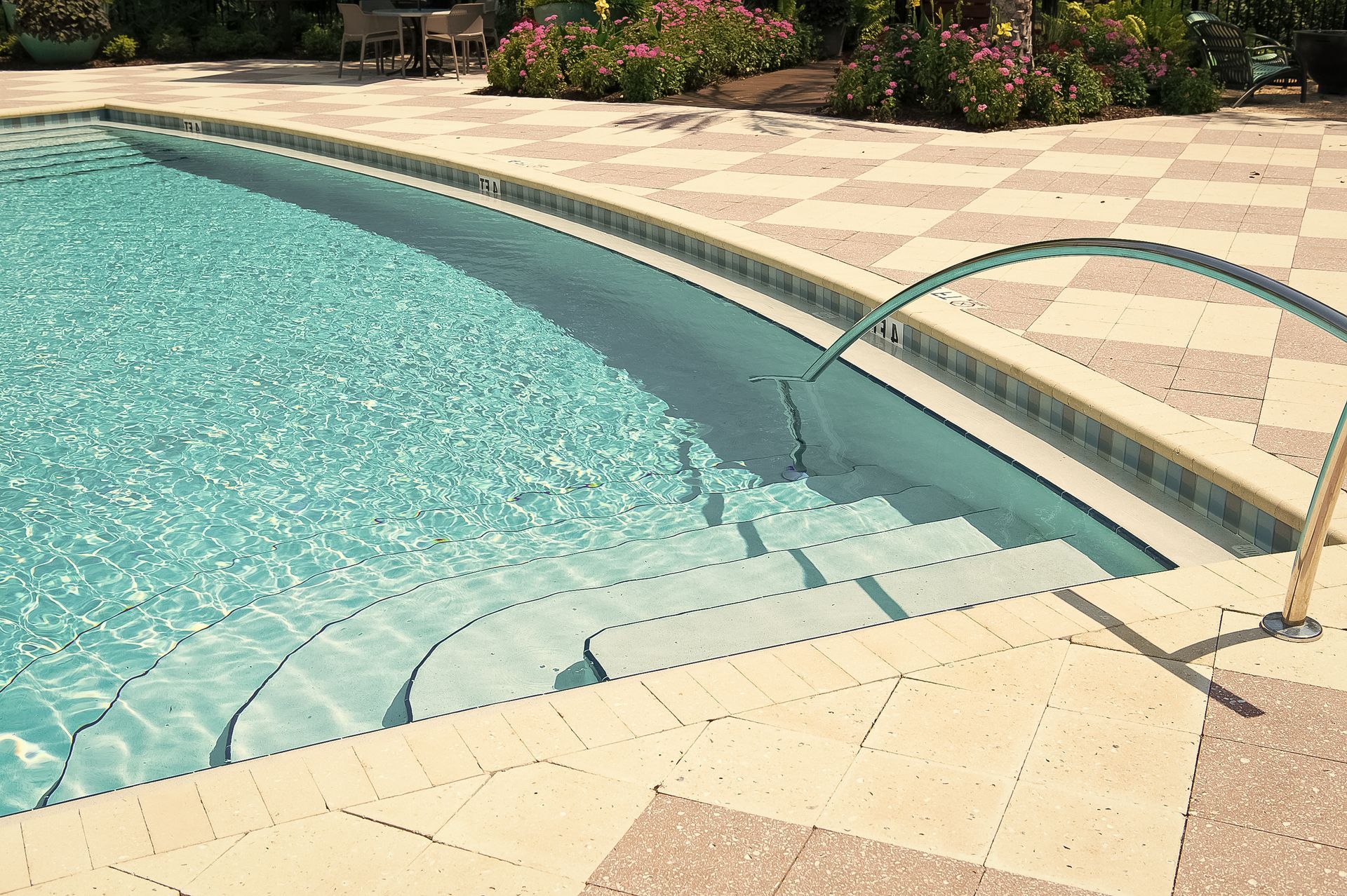 Curved swimming pool with steps, metal handrail, and tan tile deck in bright sunlight