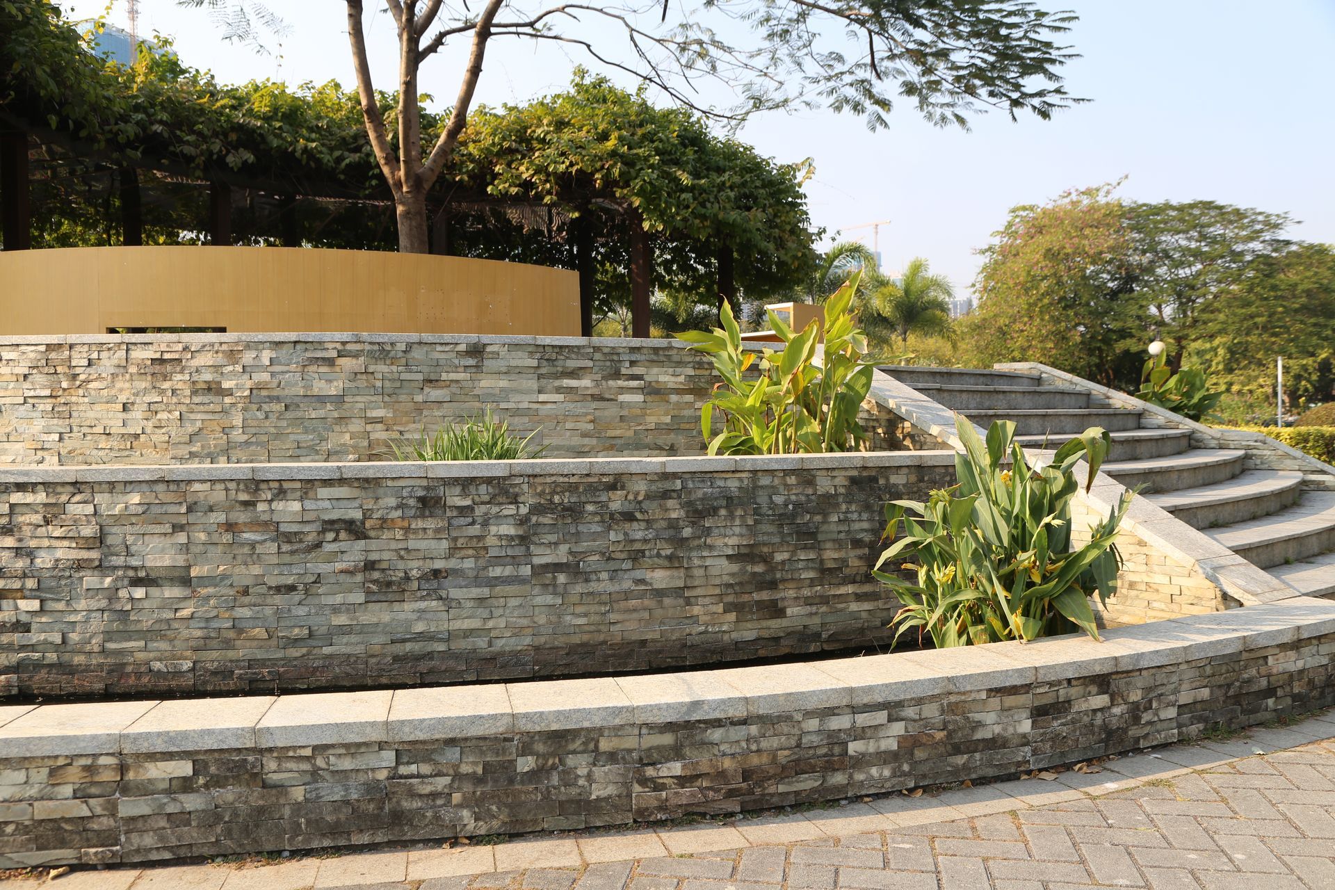 Terraced stone garden wall with steps, green plants, and trees in a sunny outdoor park setting