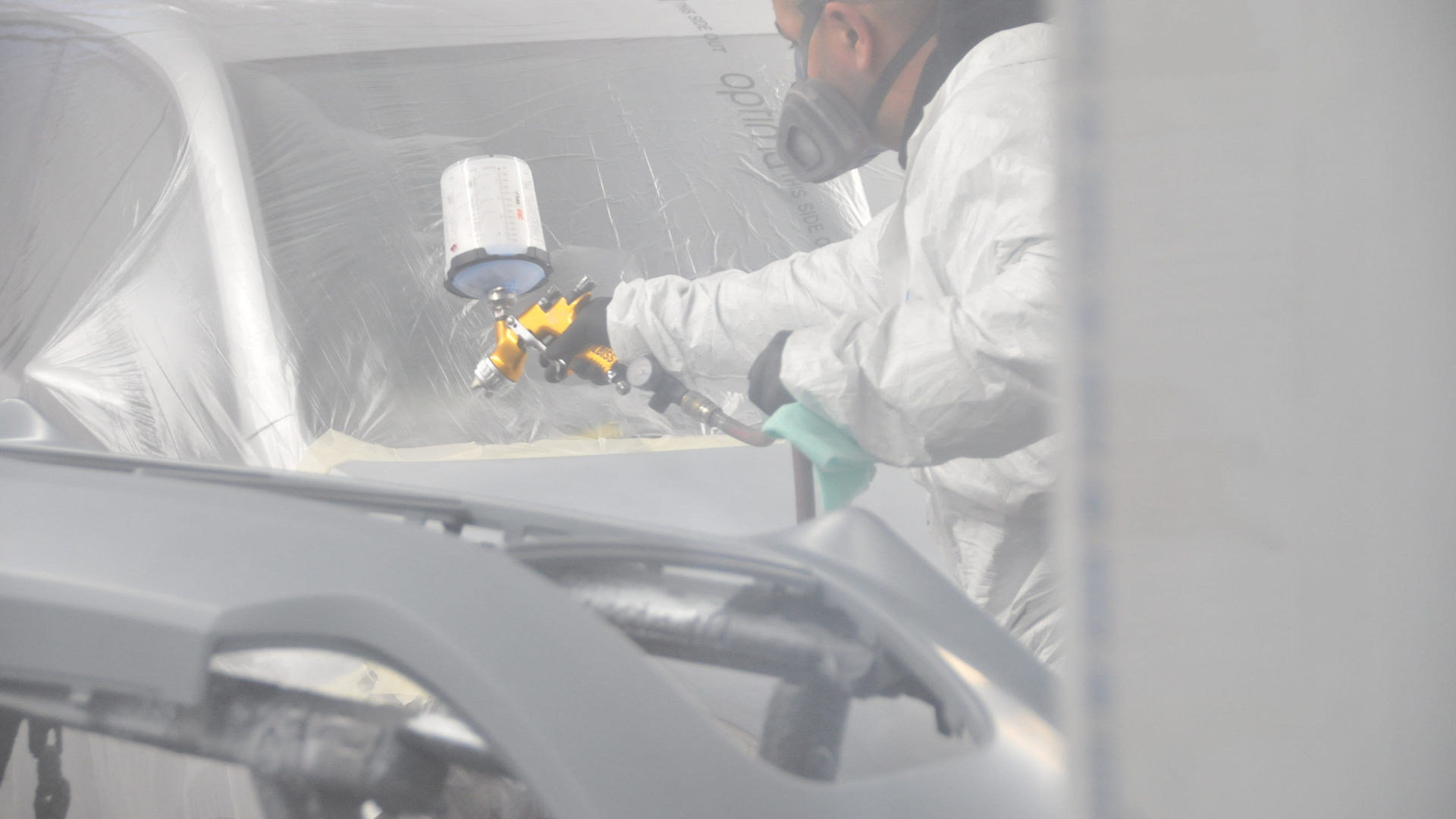 A person in protective gear sprays paint onto a car part in a spray booth.
