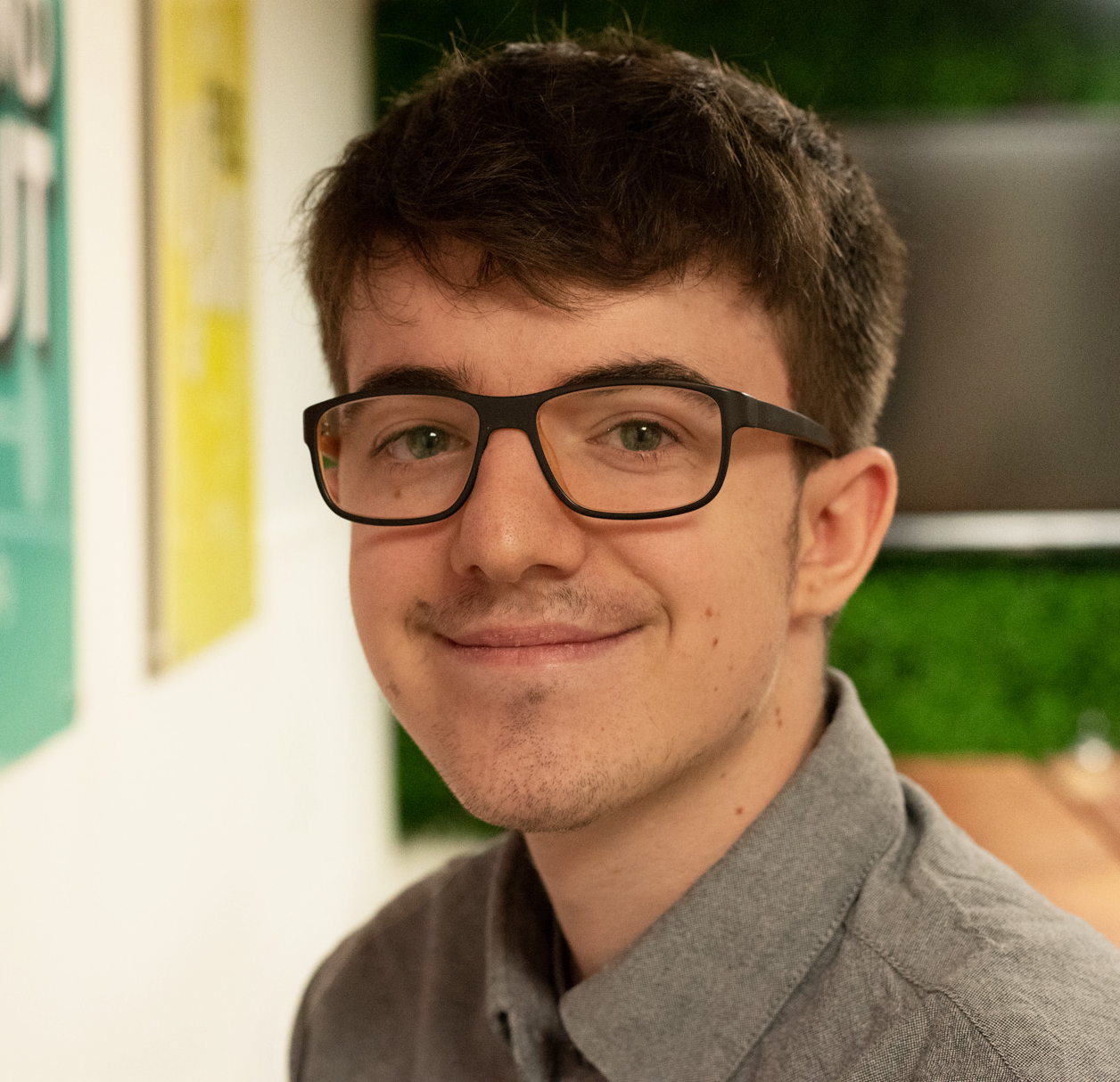 A person with short brown hair and dark-rimmed glasses smiling at the camera in a brightly lit indoor space.