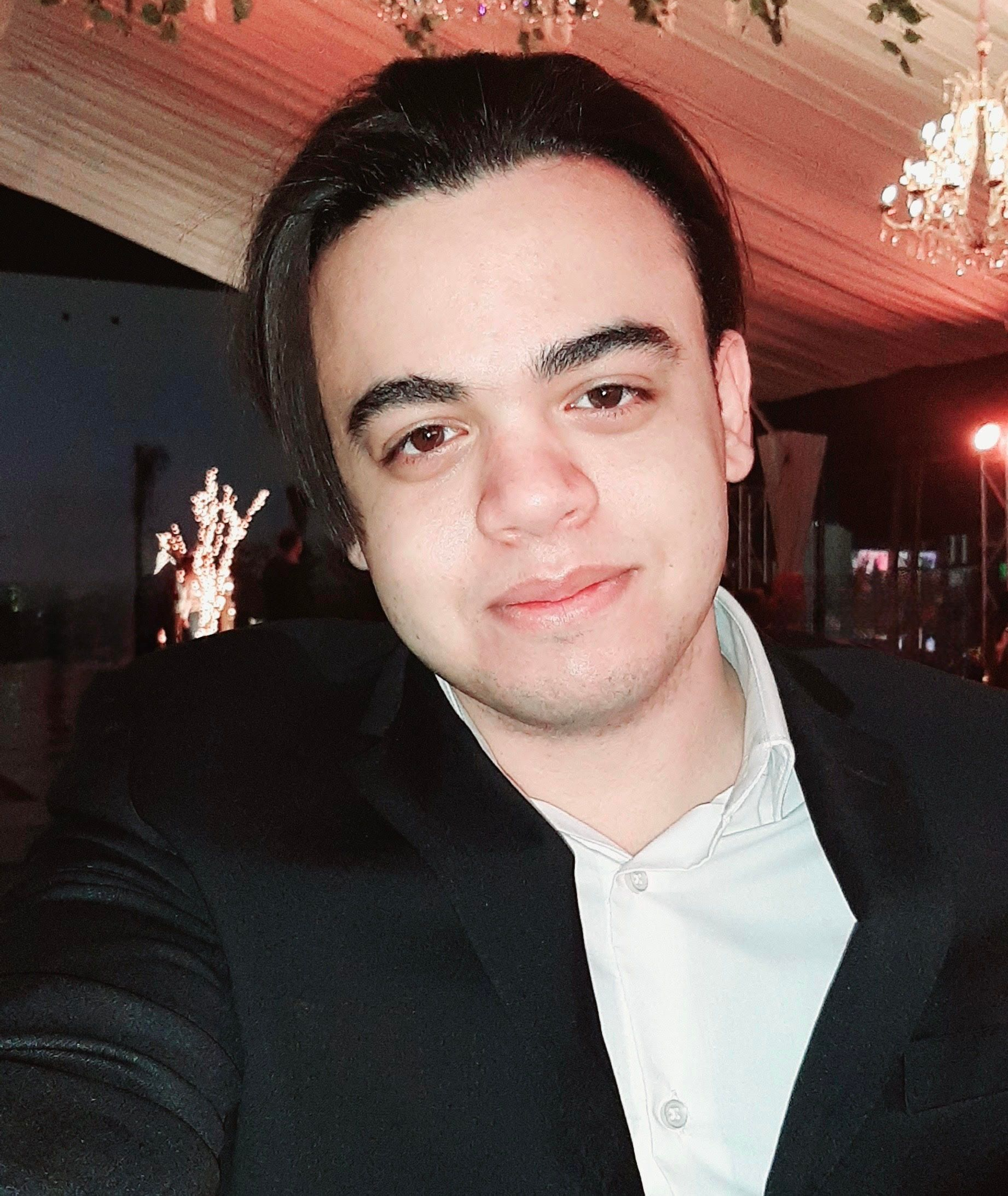 A person with dark hair wears a white button-down shirt and a black blazer, posing in an outdoor evening setting.