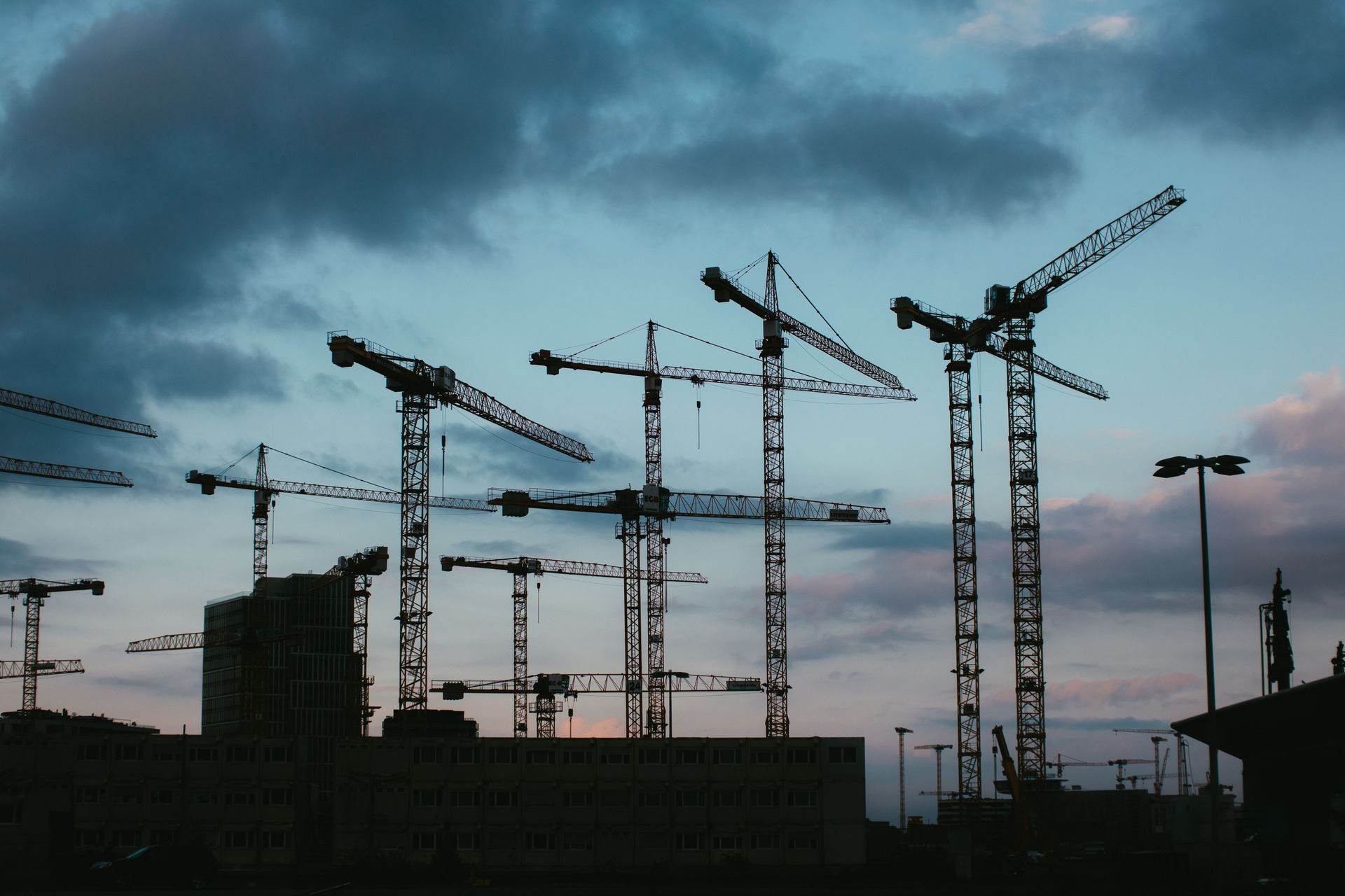 Silhouetted construction cranes at dusk against a cloudy sky over a building site